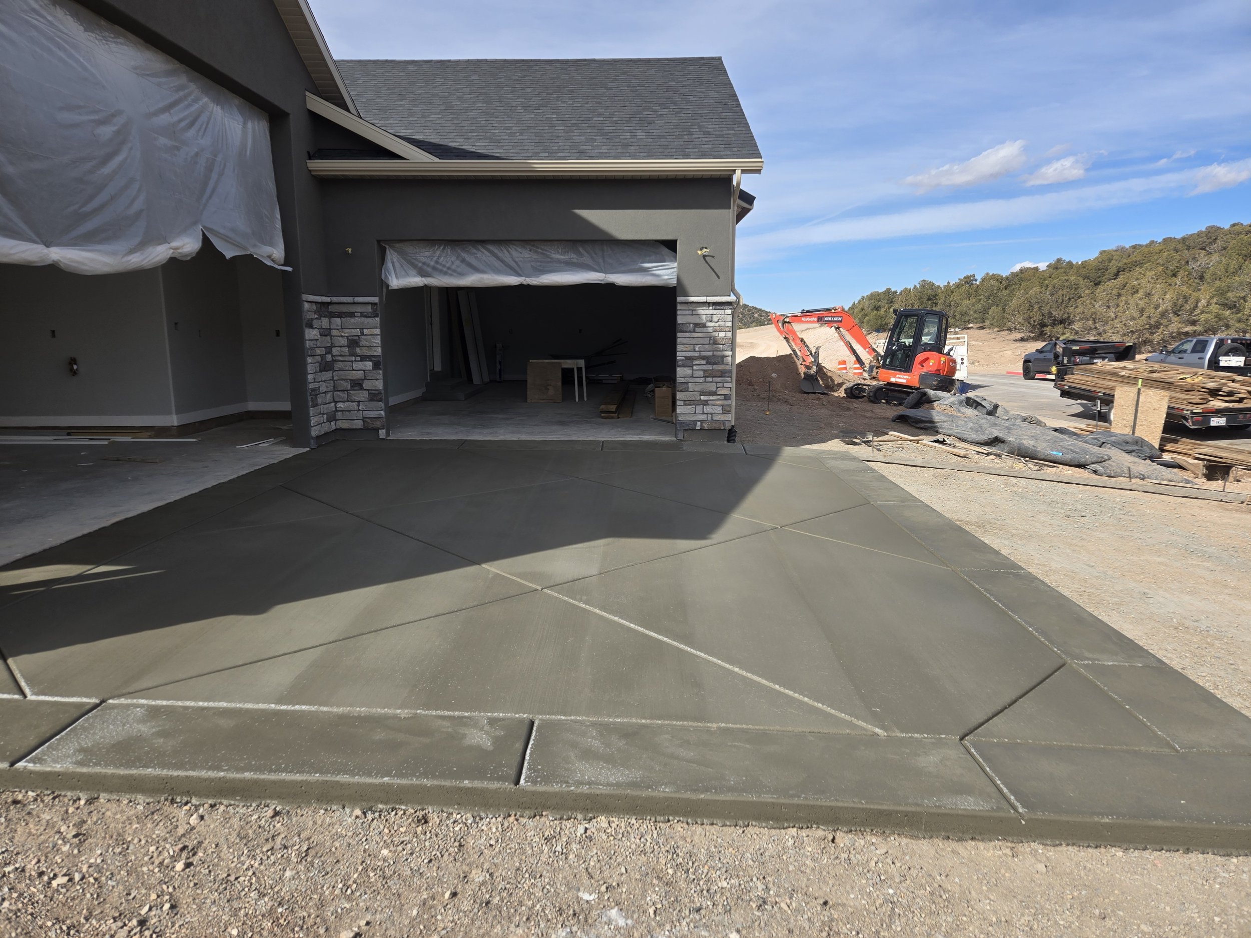 Partially constructed house with a gray exterior, stone accents, and an open garage. Construction materials and equipment are visible, and the driveway is freshly poured concrete. Construction is ongoing with a mini excavator and other building suppl