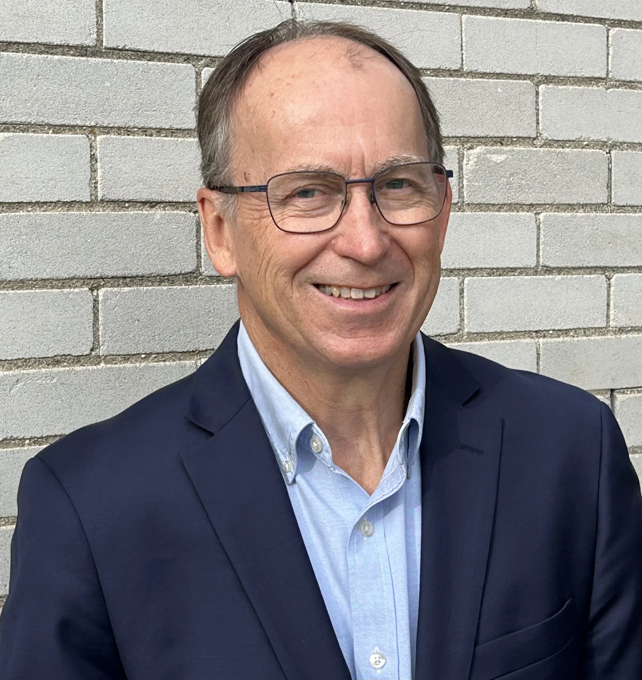 A smiling middle-aged man with glasses, wearing a navy blue suit jacket and a light blue button-up shirt, standing in front of a white brick wall.