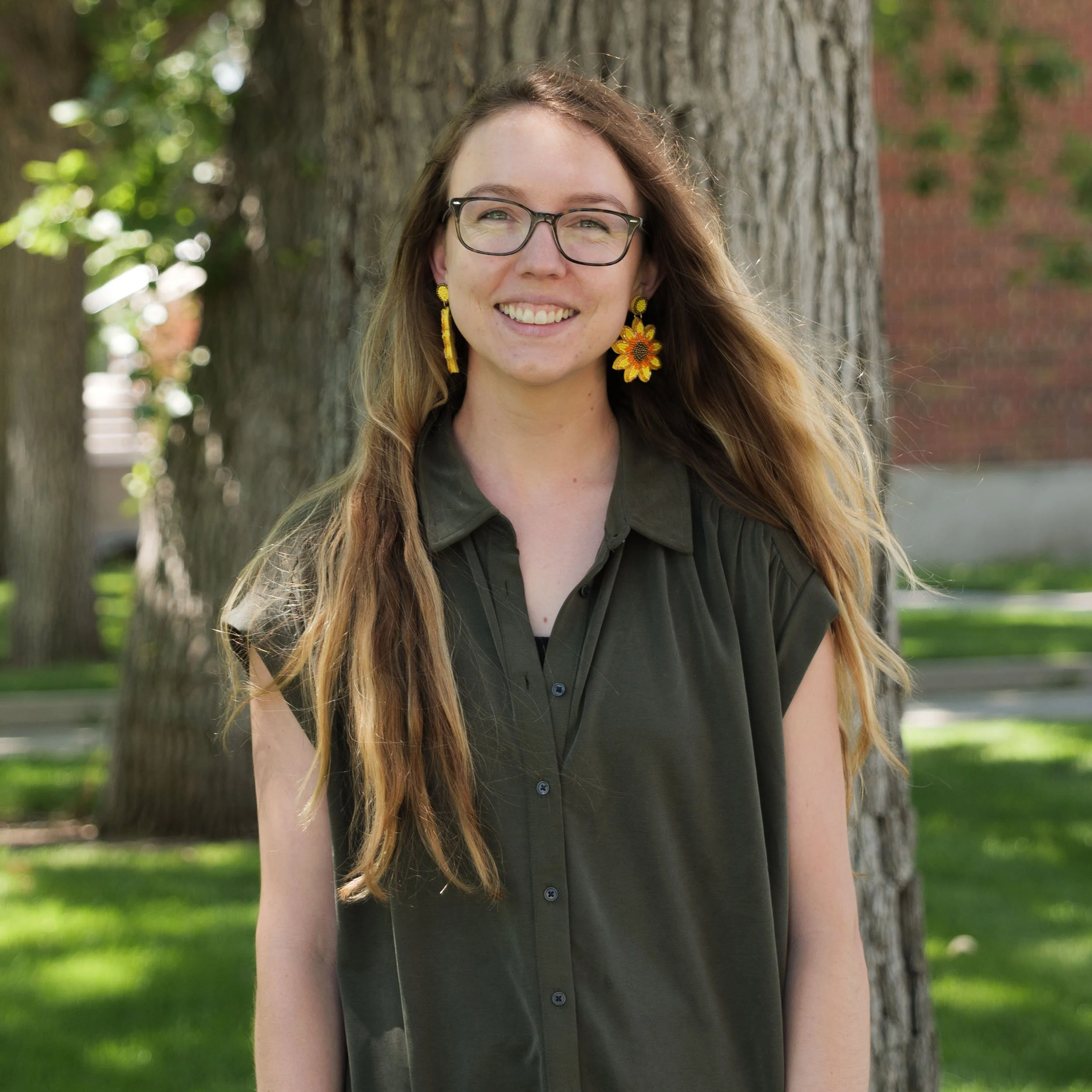 Young woman with long hair, glasses, and colorful earrings outdoors near a tree, smiling.