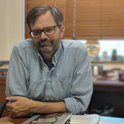 A man with glasses and a beard, wearing a light blue shirt, sits at a desk in an office environment with wooden blinds and shelves in the background.