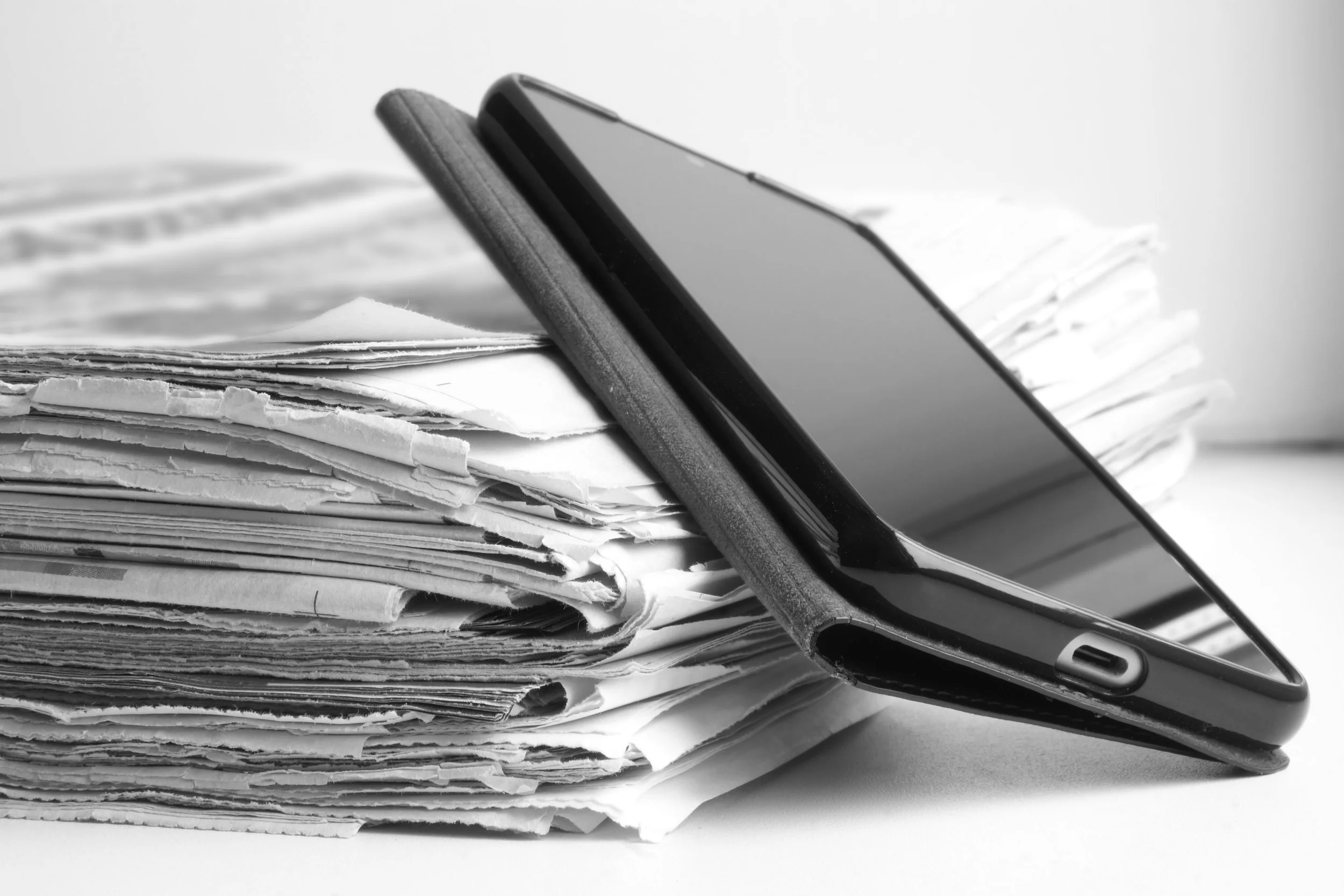 Black and white photo of a smartphone resting on top of a large stack of disorganized papers and documents.