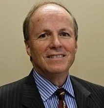 A man with short, light-colored hair wearing a dark suit, blue striped shirt, and a tie, smiling in front of a plain beige background.
