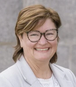 Portrait of a middle-aged woman with short, light brown hair, wearing glasses and a white blazer, smiling against a neutral background.