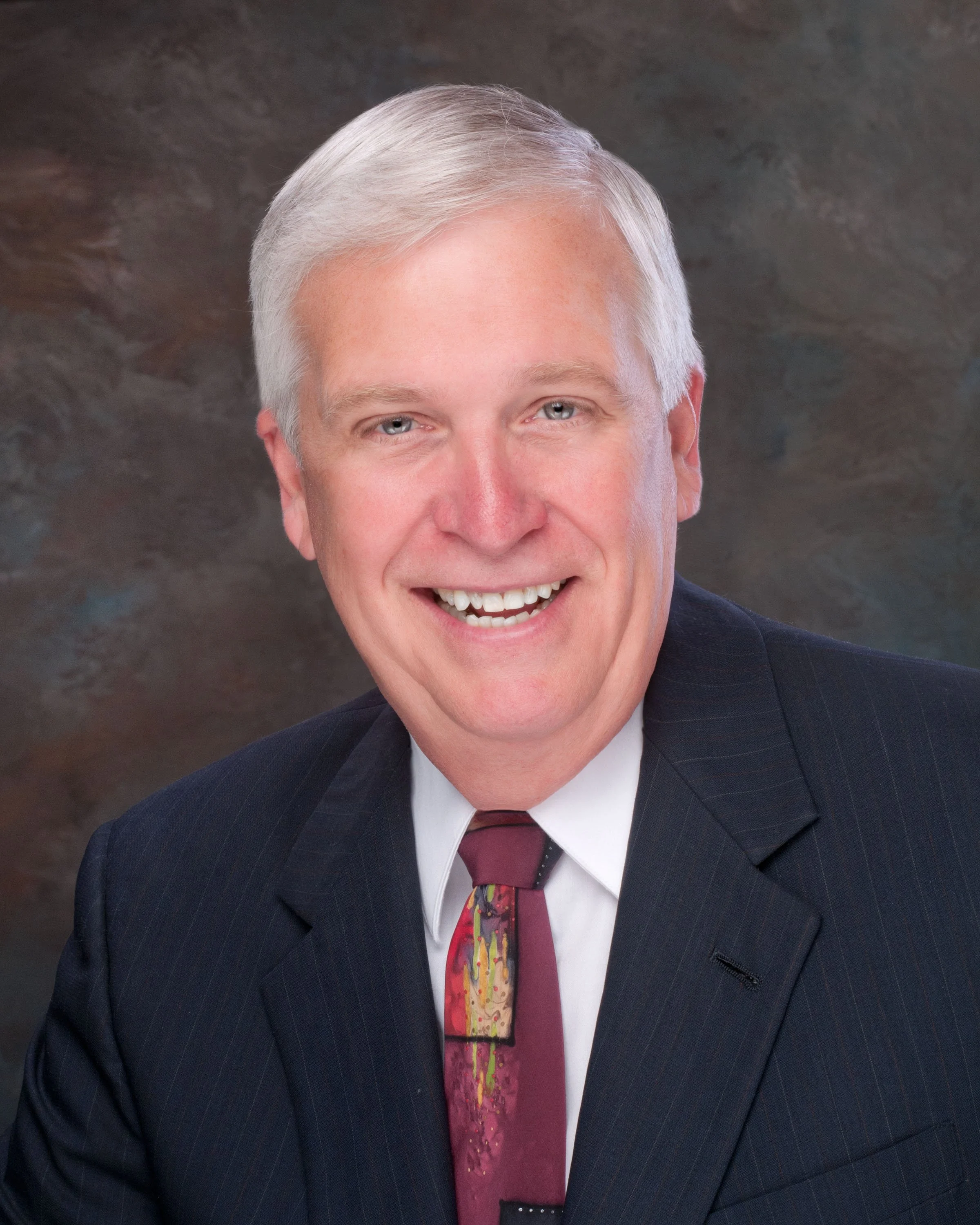 A smiling man with gray hair wearing a dark suit, white shirt, and a colorful tie in front of a textured background.