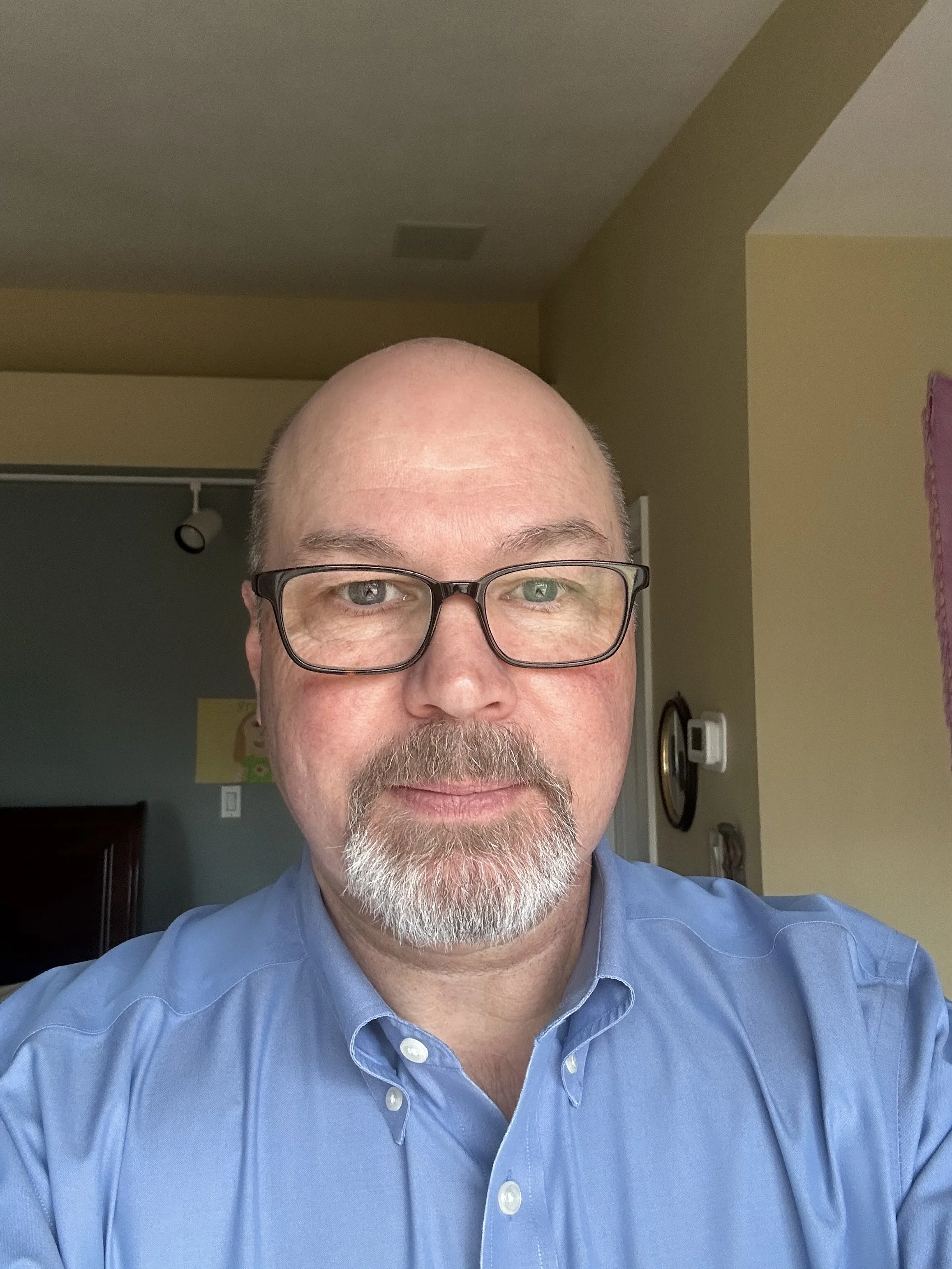 A man with glasses, a bald head, and a gray beard takes a selfie indoors, wearing a blue collared shirt, with a background that includes a hallway with a thermostat and a partial view of a doorway.