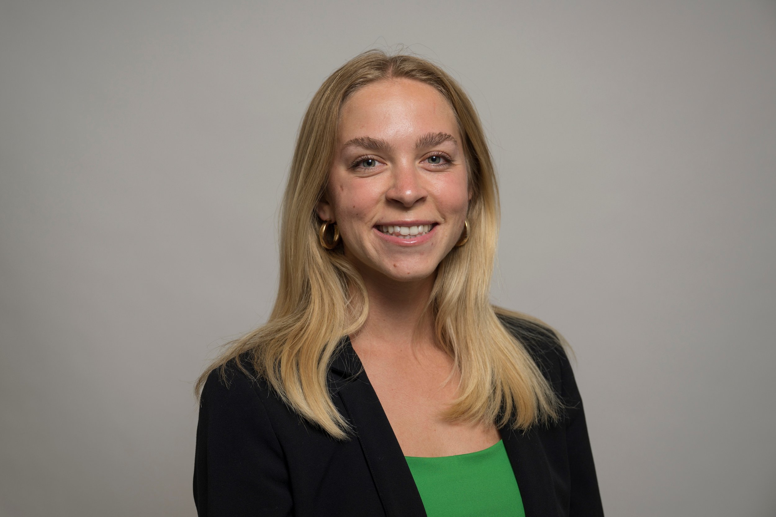 A smiling woman with long blonde hair, gold hoop earrings, wearing a black blazer over a green top, standing against a plain light gray background.
