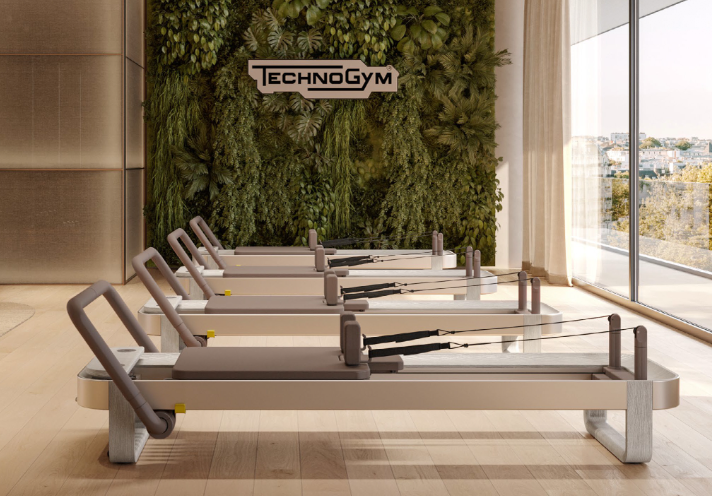 Five Pilates reformer machines lined up in a fitness studio with a green living wall and a 'TechnoGym' sign, next to large windows with a city view.
