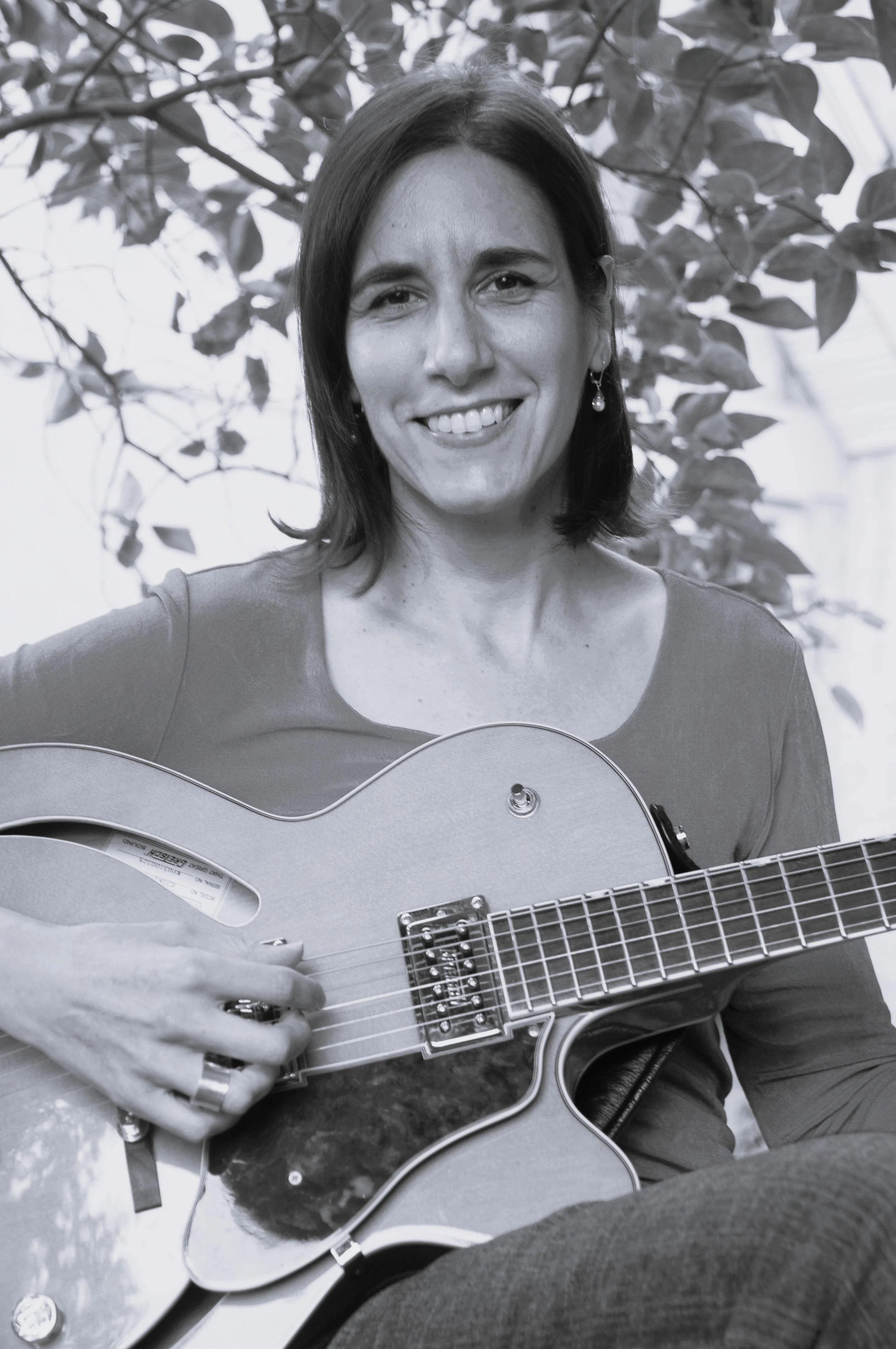 Leah R.G. is smiling while playing an acoustic guitar outdoors, with leafy branches in the background.