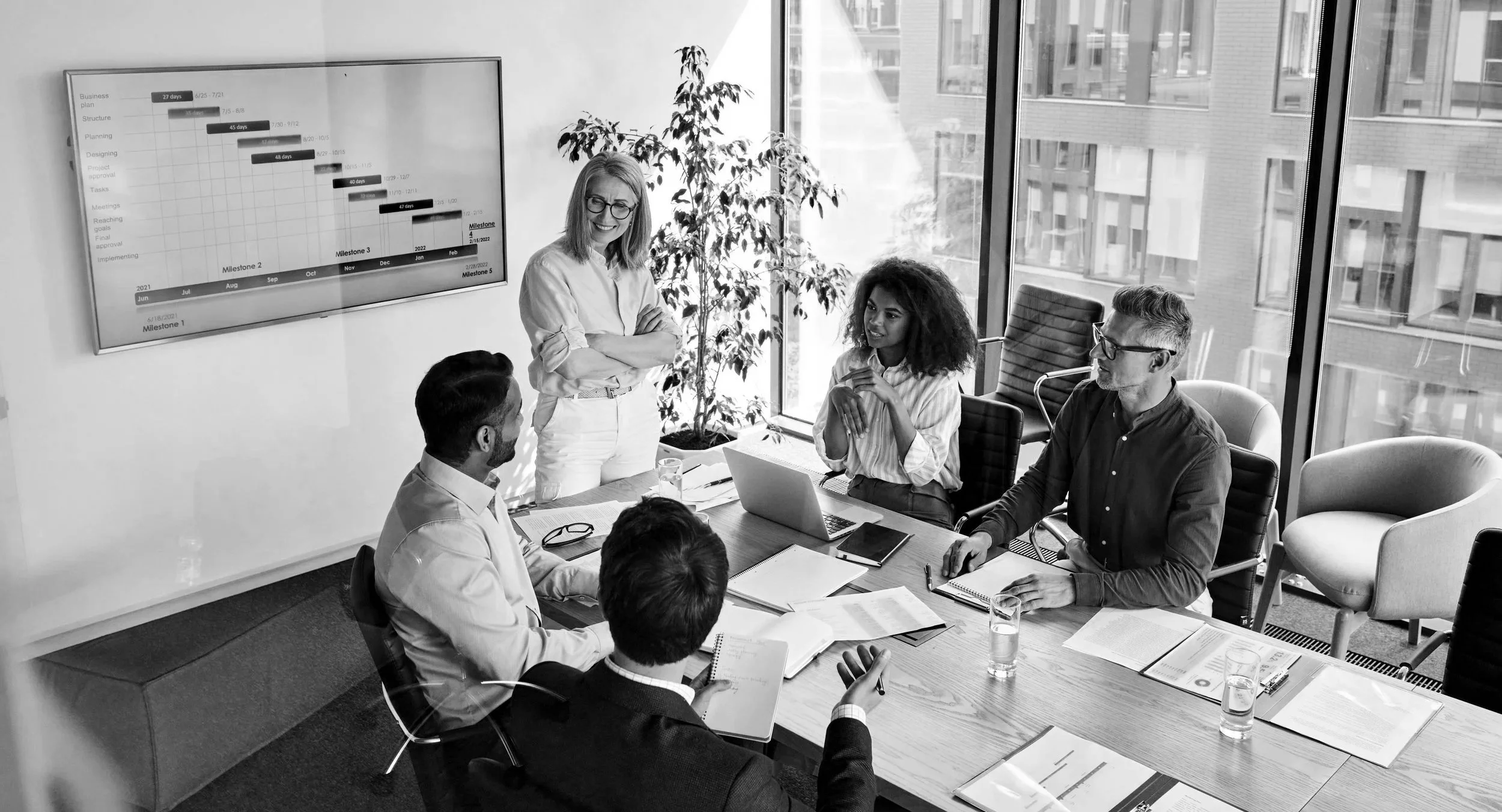 A group of six diverse professionals in a modern office meeting room with large windows, gathered around a rectangular table with documents and glasses of water, engaged in a discussion. One woman is standing near a large screen displaying a Gantt chart, smiling as she talks to the seated colleagues.