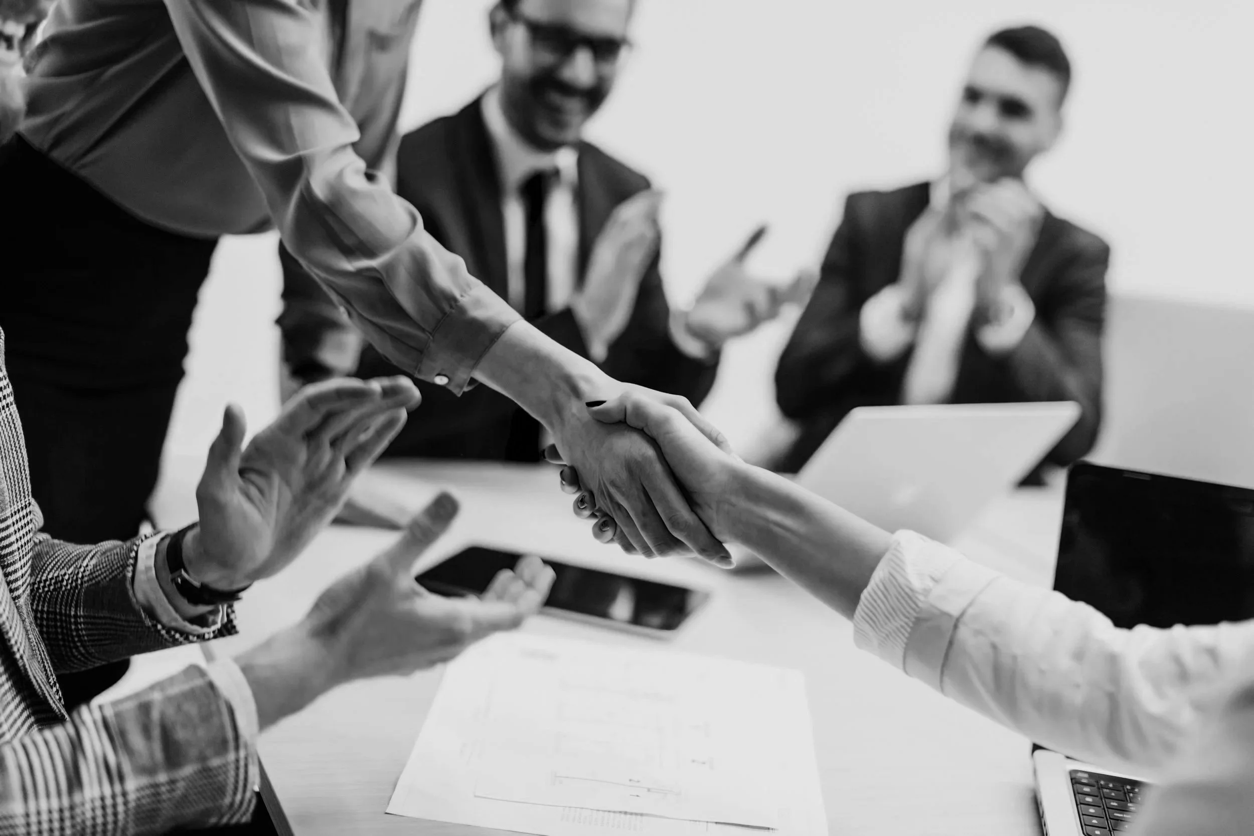 People in a business meeting, with one person shaking hands and others clapping or holding phones and laptops.