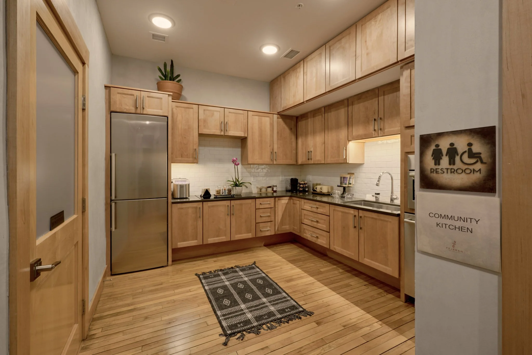 A community kitchen with wooden cabinets, a refrigerator, a sink, potted plant, and decorative items on the counter, with a sign indicating restrooms.