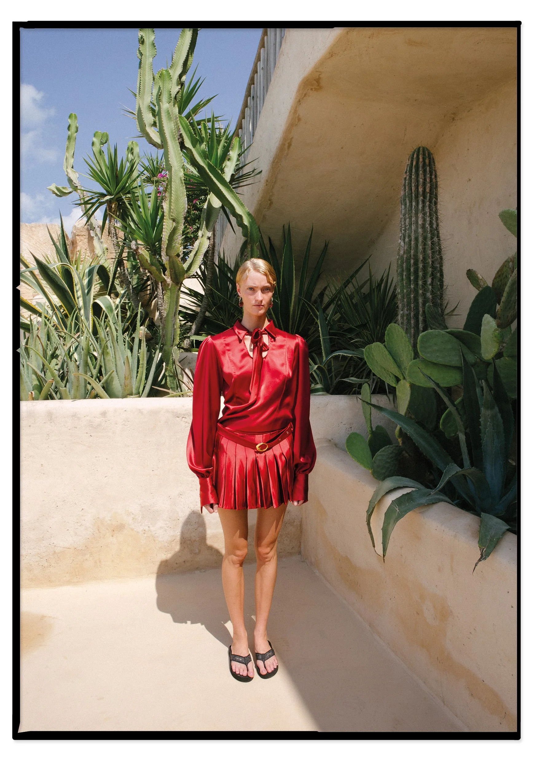 Woman in a red shiny blouse and pleated mini skirt standing outdoors among large green cacti and succulents with a sunny sky above.