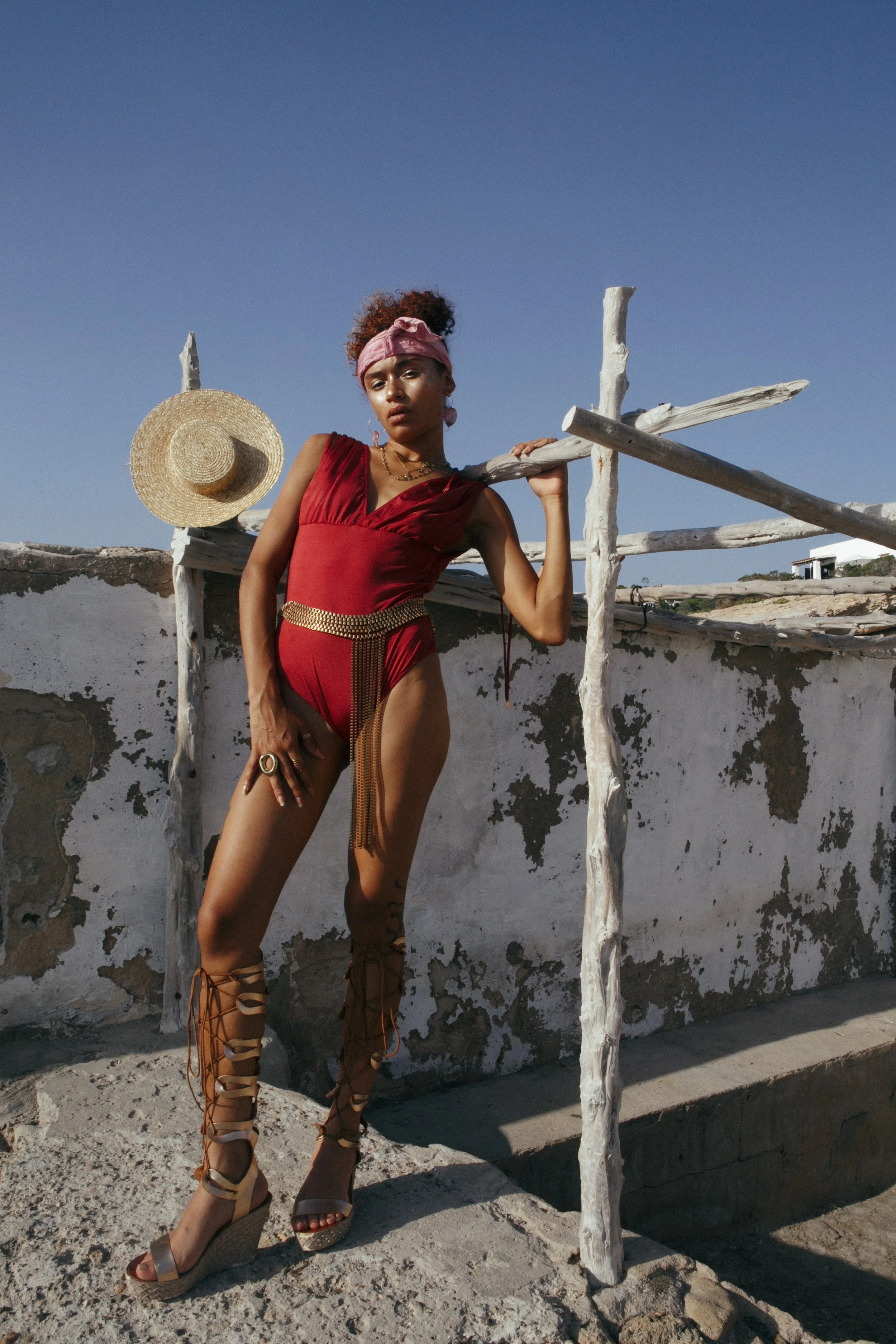 A woman in a red swimsuit with gold accessories and lace-up wedge heels poses on an outdoor rooftop, holding a wooden staff with a rustic background and clear blue sky.