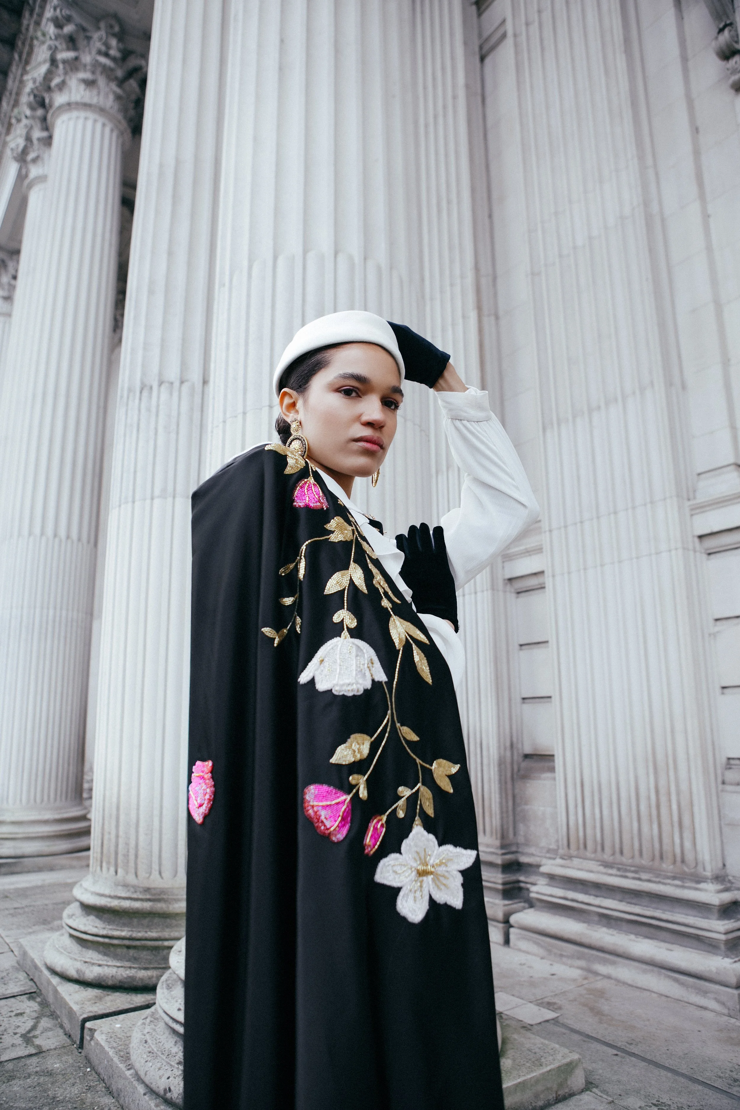 A woman standing near large white classical columns, wearing a black embroidered cape with floral designs, a white shirt, black gloves, and a white beret, posing confidently.