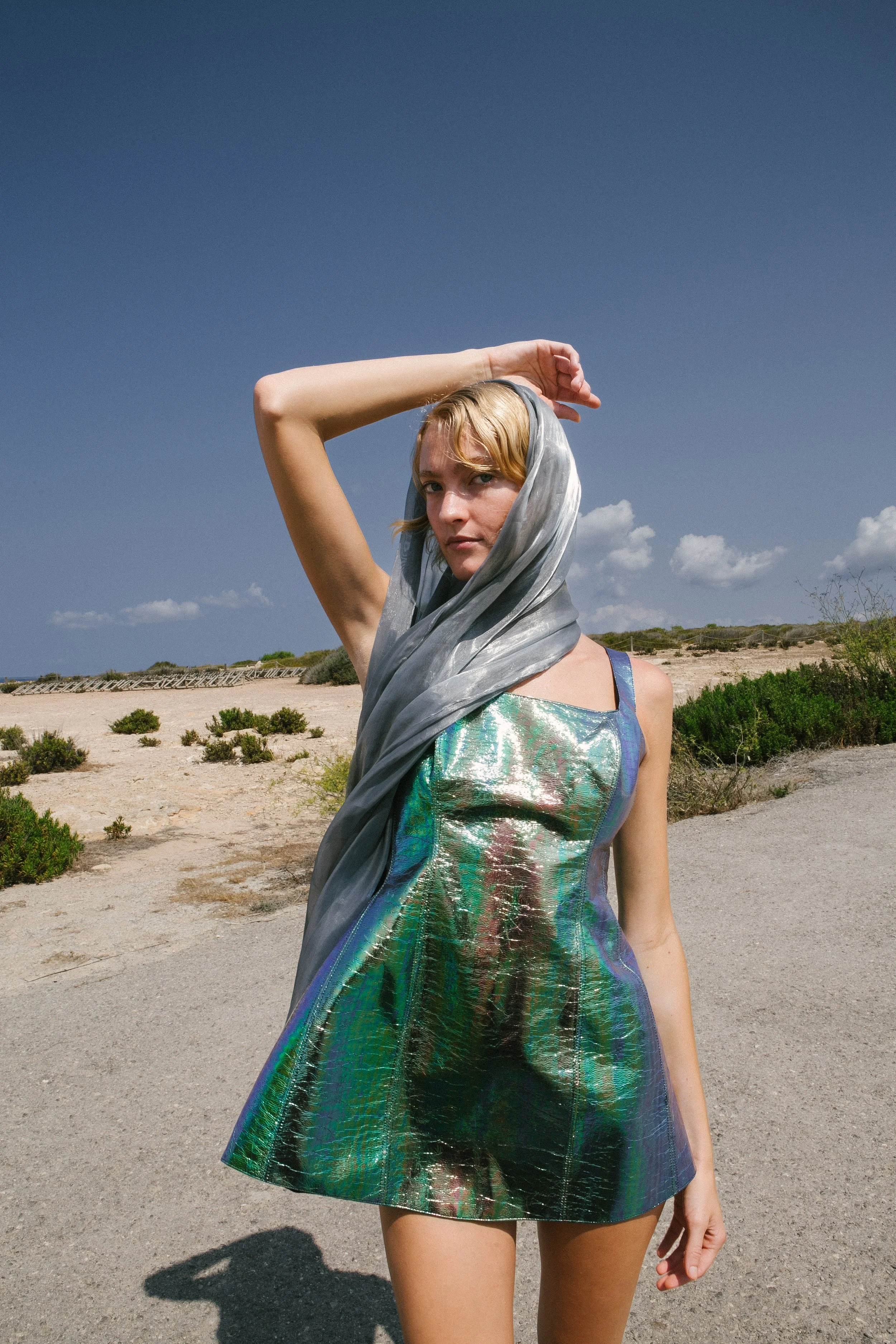 A woman standing outdoors in a desert-like area with shrubs and a clear blue sky, wearing a shiny iridescent dress and a gray scarf over her head.