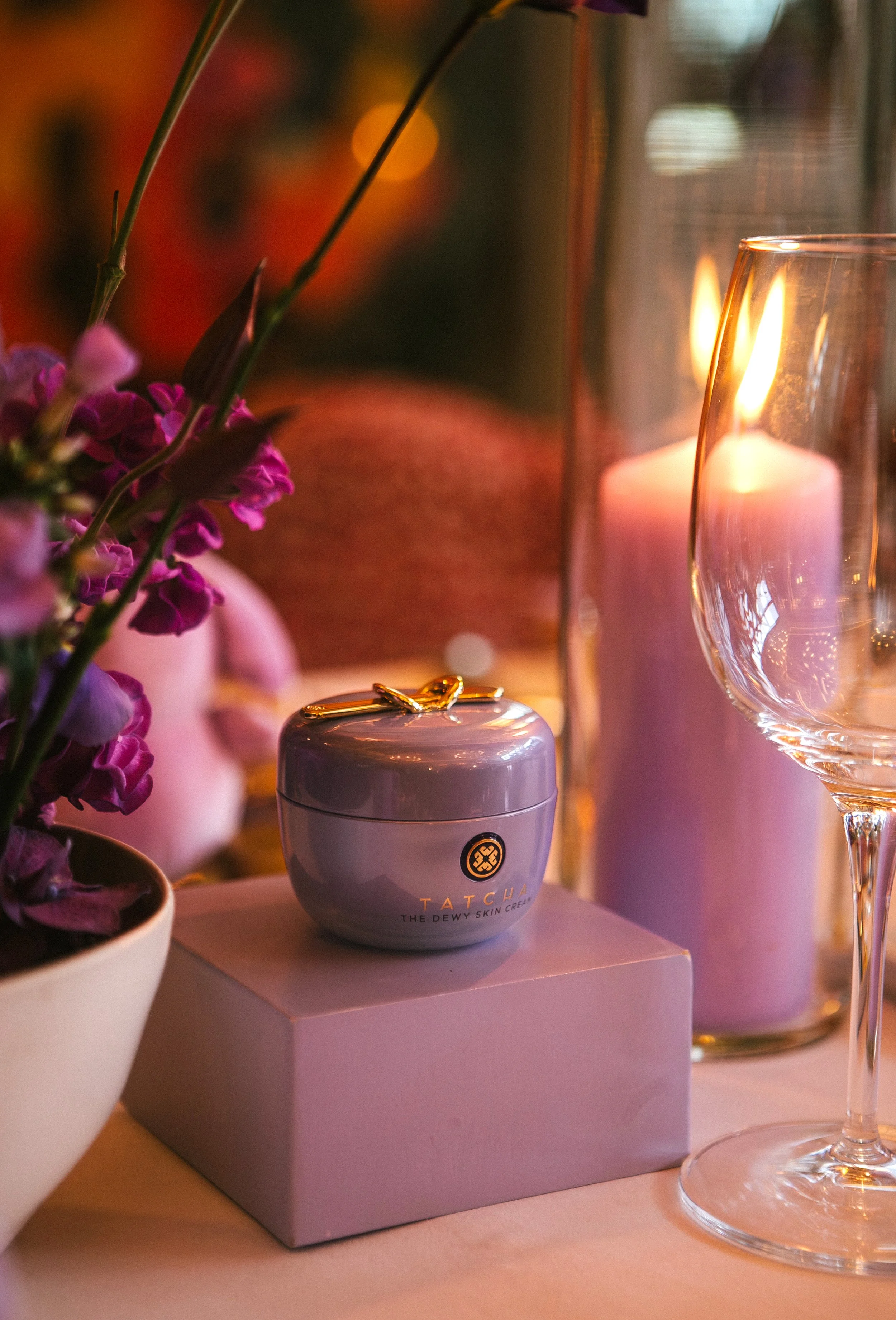 A small jar of Tatcha skincare cream with a gold clasp on top, placed on a pink box. Surrounding elements include purple flowers in a white vase, lit candles, and a wine glass, all with warm ambient lighting.