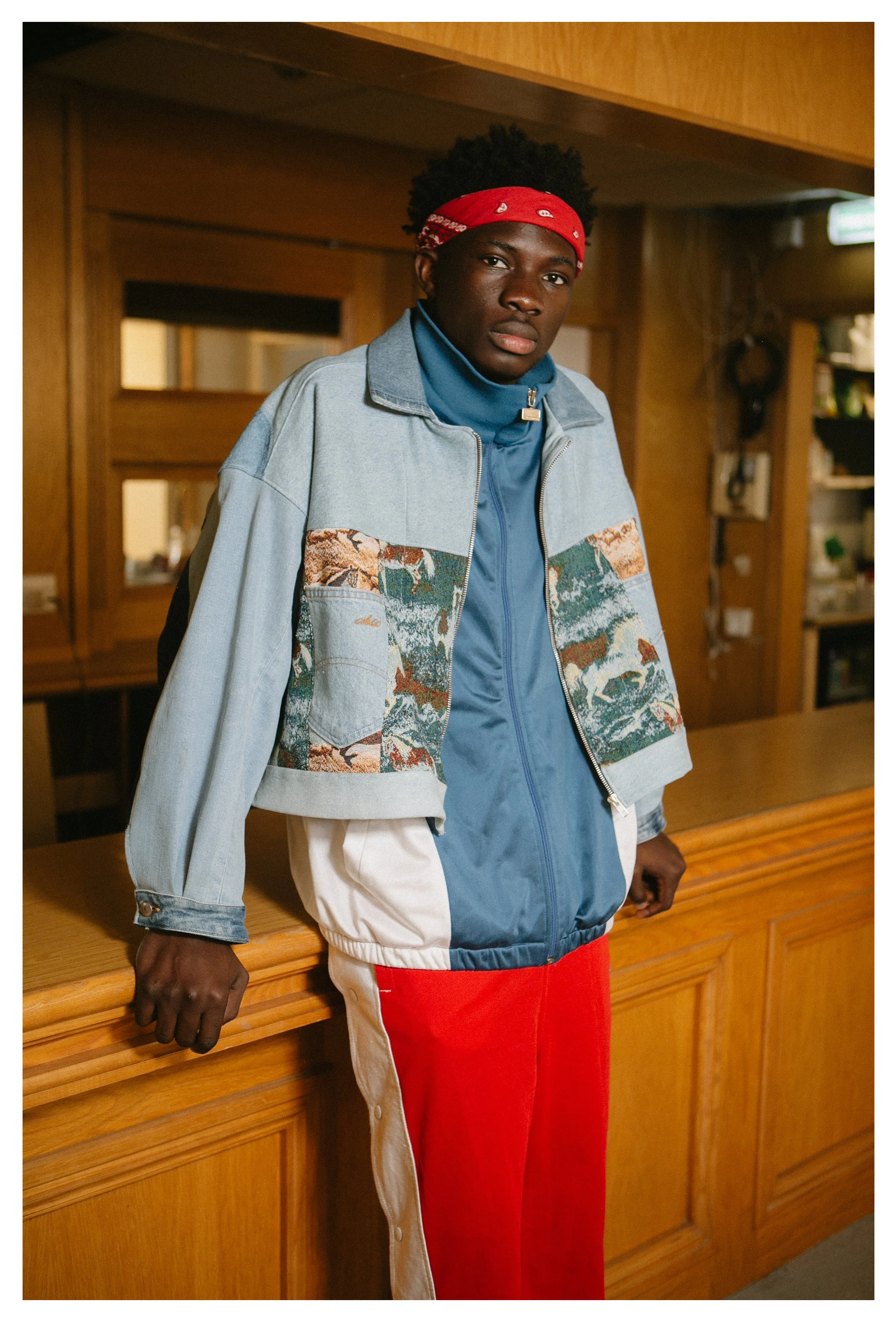 A young man with dark skin, curly black hair, wearing a red bandana, stands indoors at a wooden counter, wearing a light jeans jacket with a landscape pattern on the chest, a blue zip-up jacket underneath, and red pants with white side stripes.