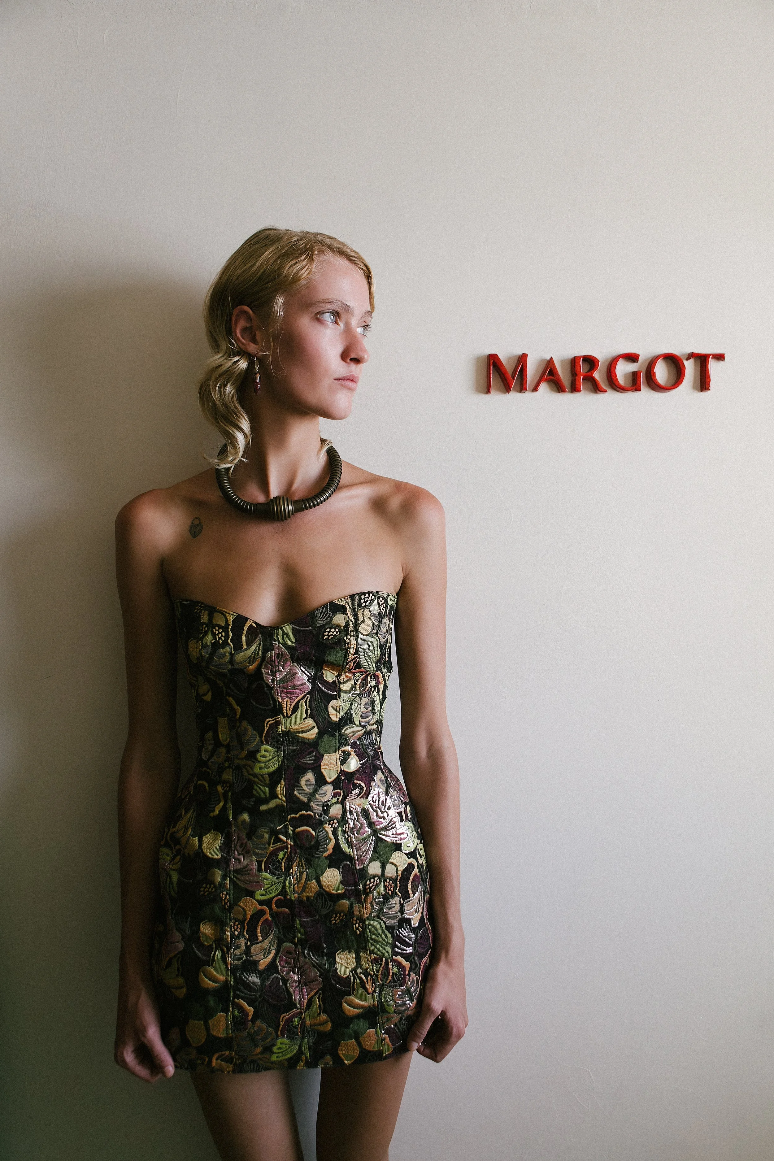 A woman with blonde hair standing against a beige wall, looking to her left. She is wearing a strapless, patterned dress with dark floral designs, a black necklace, and purple earrings. There is a small tattoo on her left shoulder. Red letters spelli