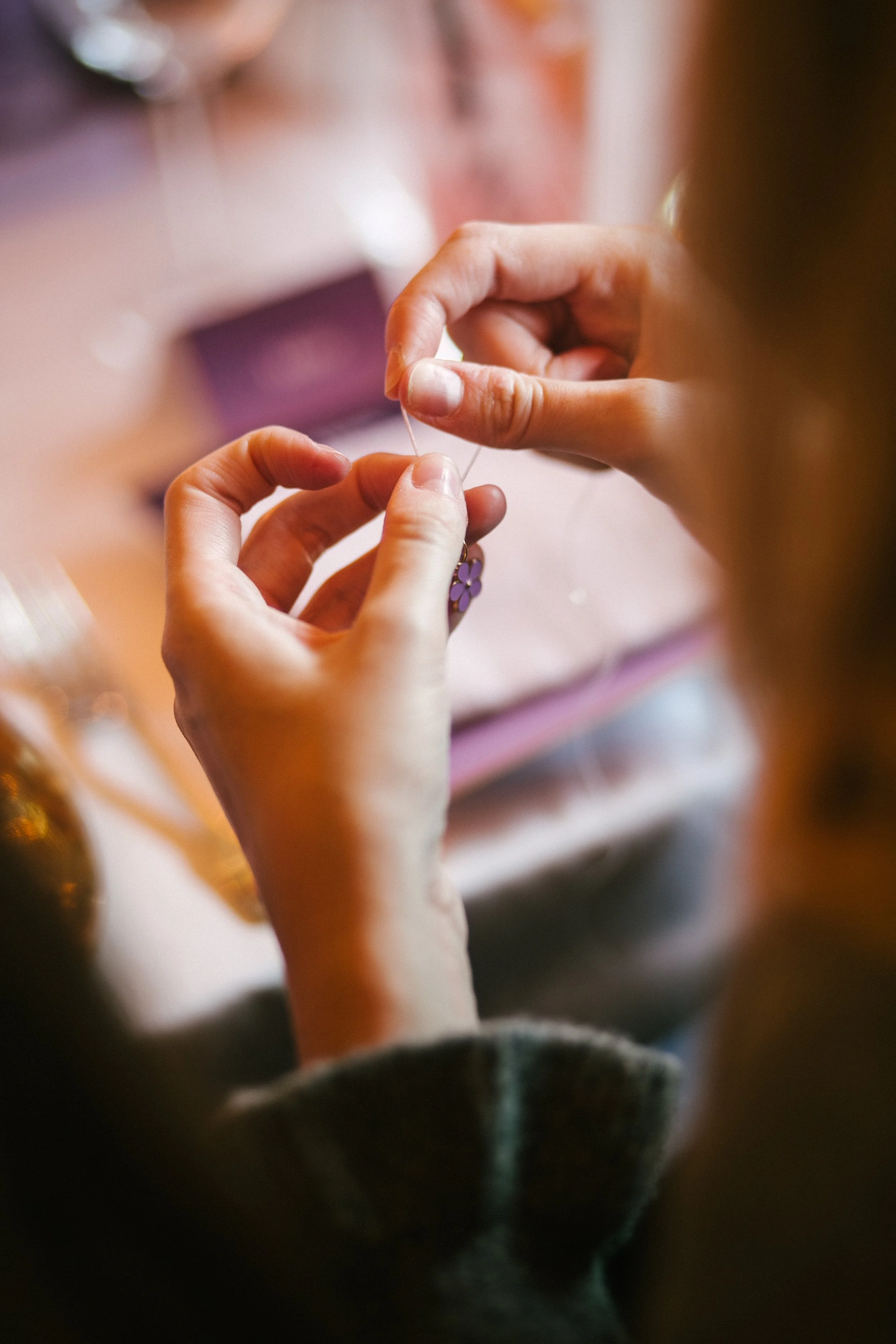 A person is threading a purple flower-shaped charm onto a string or wire, with a softly blurred background of a table and various objects.