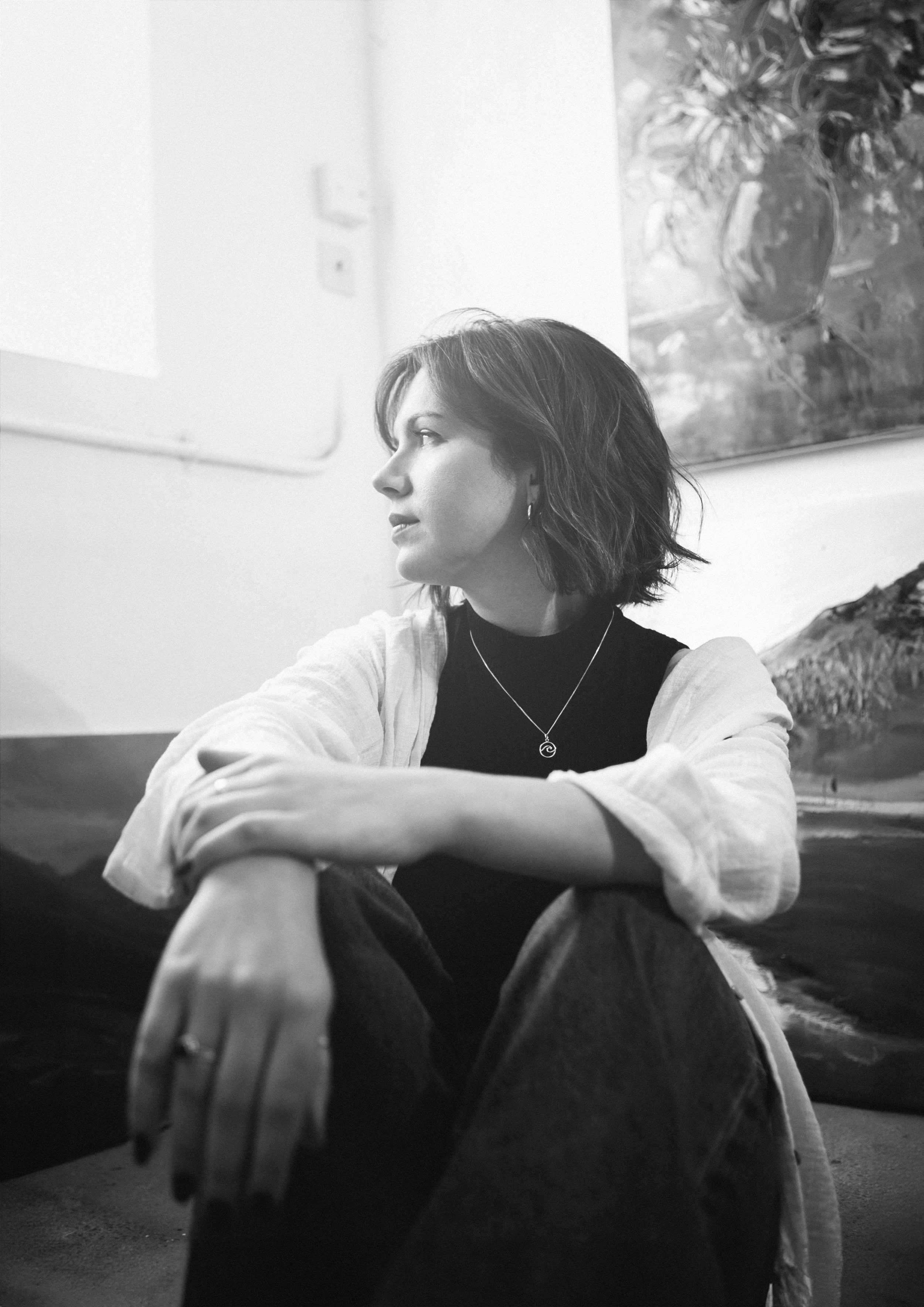 Black and white photo of a woman with short hair, sitting on the floor with her arms around her knees, looking to the side. She is wearing a necklace, earrings, a dark top, and a light-colored shirt.