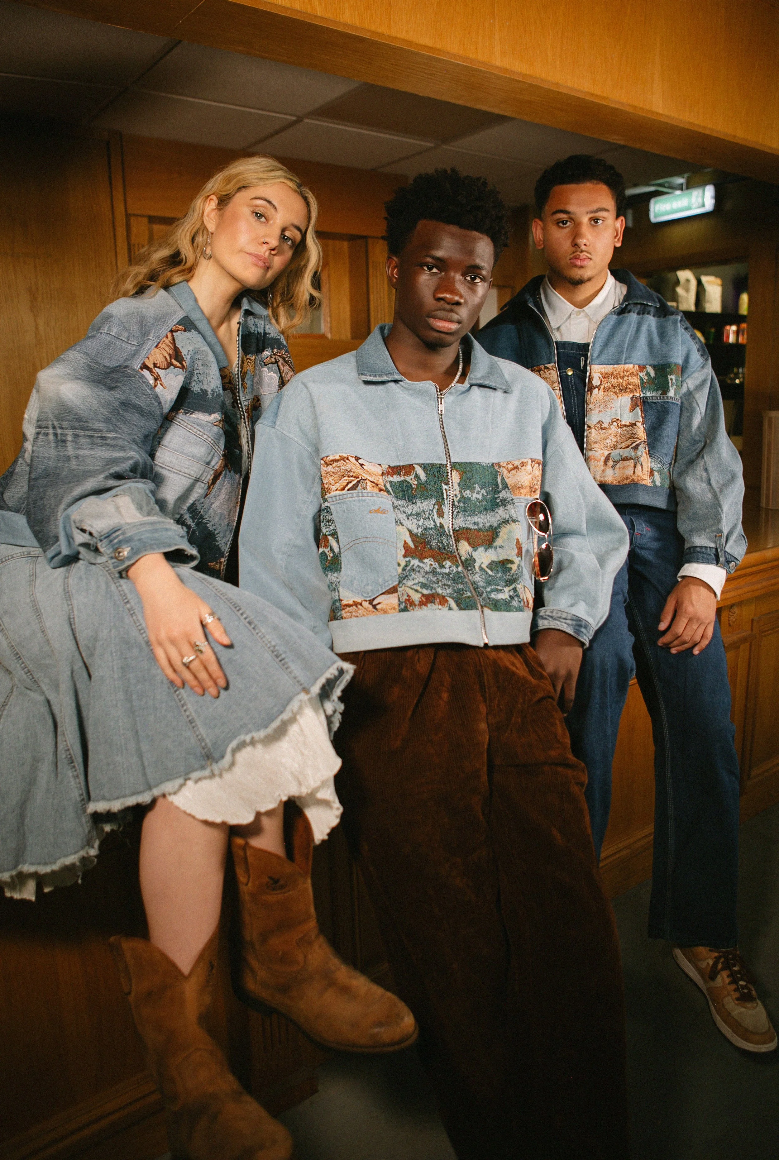 Three young adults with vintage denim jackets and casual clothing posing in a wood-paneled room.