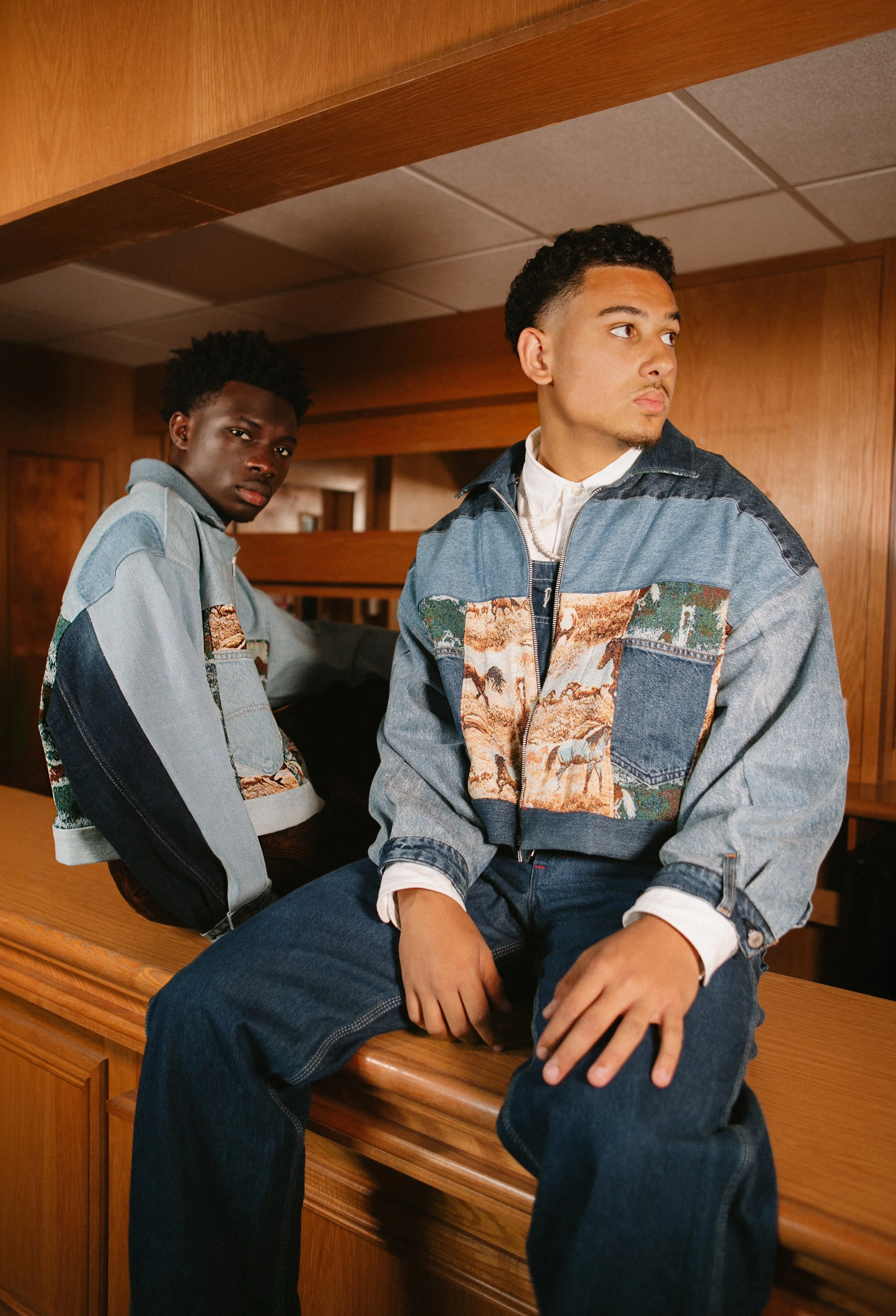 Two young men, one with curly black hair and another with short, curly hair, both wearing denim jackets with scenic landscape patches, sitting on a wooden counter in a warmly lit room with wood-paneled walls.