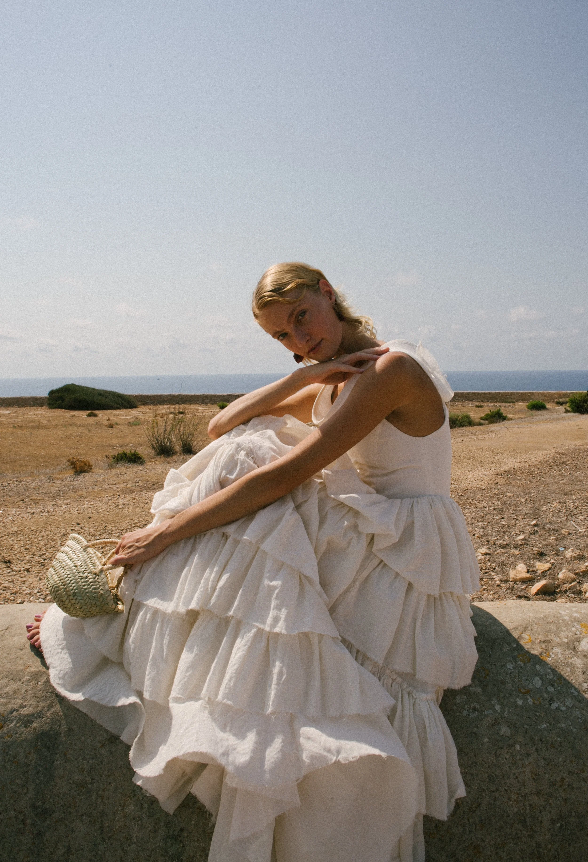 A woman in a flowing white dress sitting on a rock in a dry, open landscape with the ocean and sky in the background.