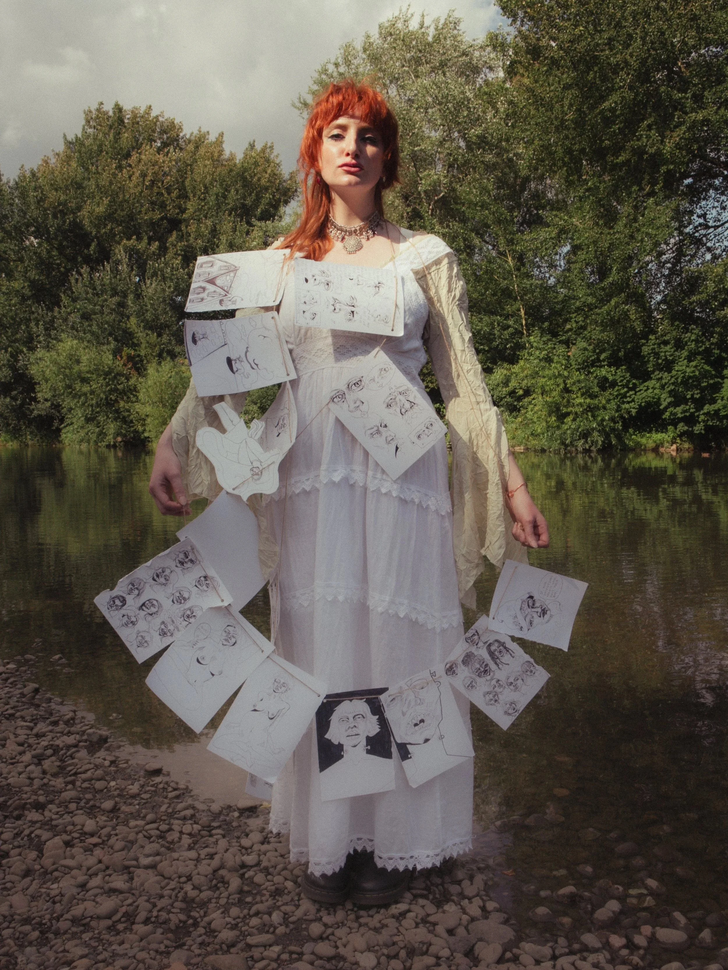 A woman with red hair wearing a white vintage-style dress and black boots standing near a river. She has sketches and drawings hanging from her shoulders, waist, and around her feet.