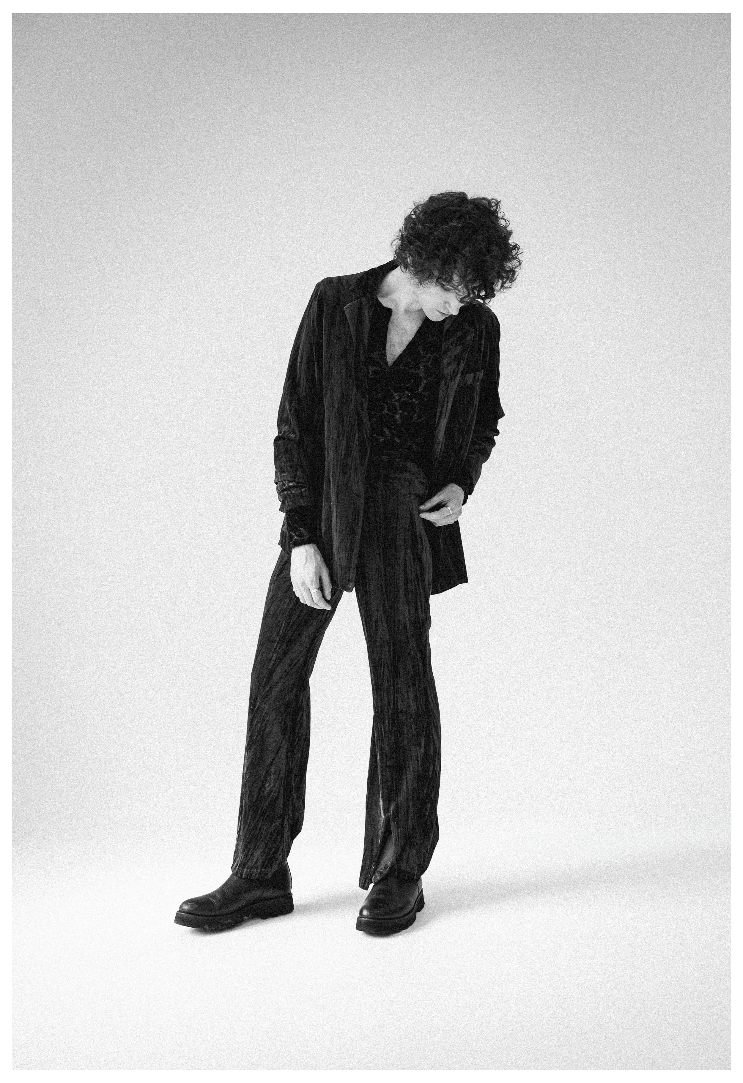 Black and white photo of a person with curly hair, wearing a dark velvety suit with a patterned shirt, standing against a plain light background, looking down and slightly to the side.