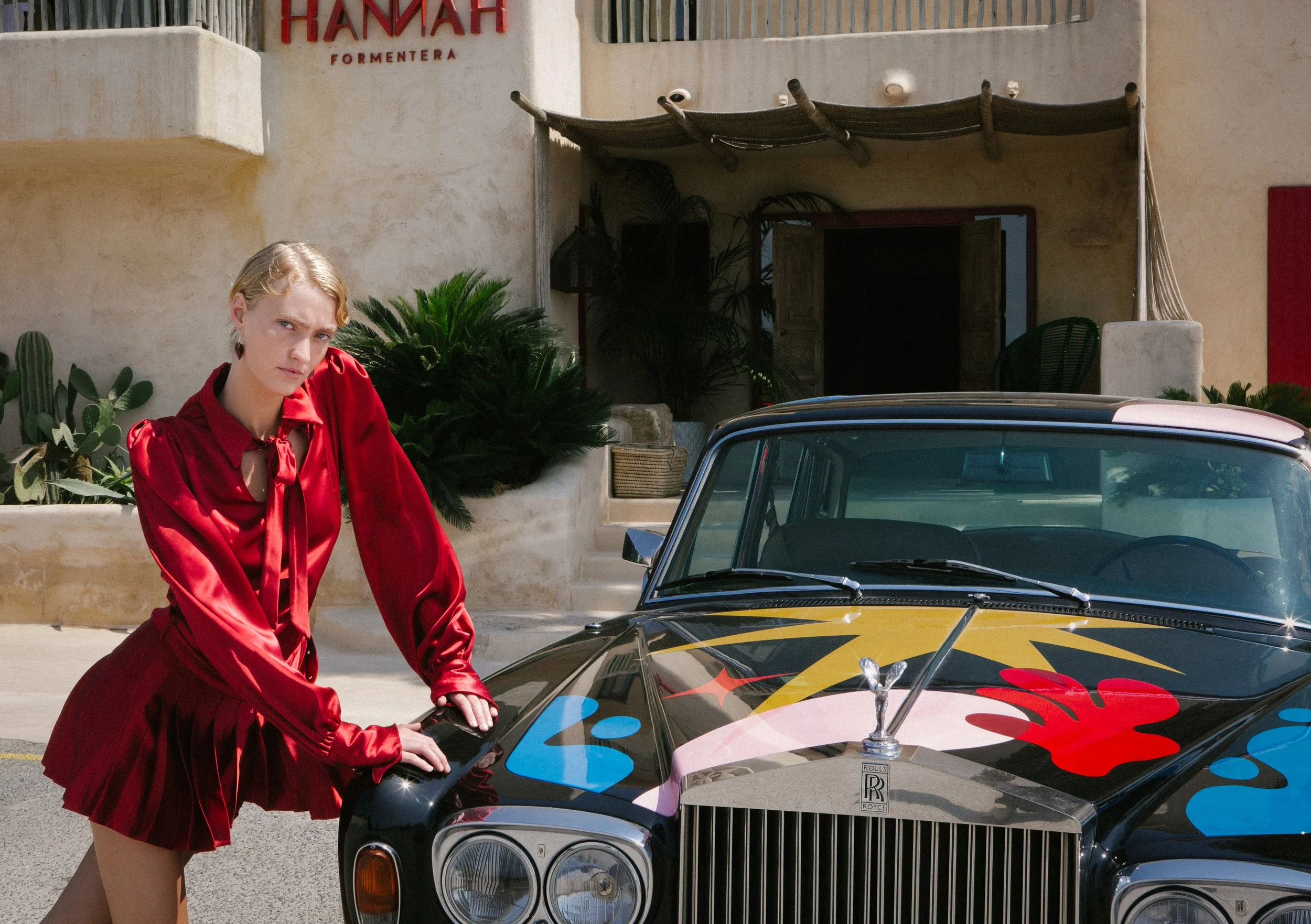 A woman in a shiny red dress leaning on a vintage black Rolls Royce car with colorful abstract paint splashes on the hood, standing outside a building with a sign that reads 'Hannah Formentera'.