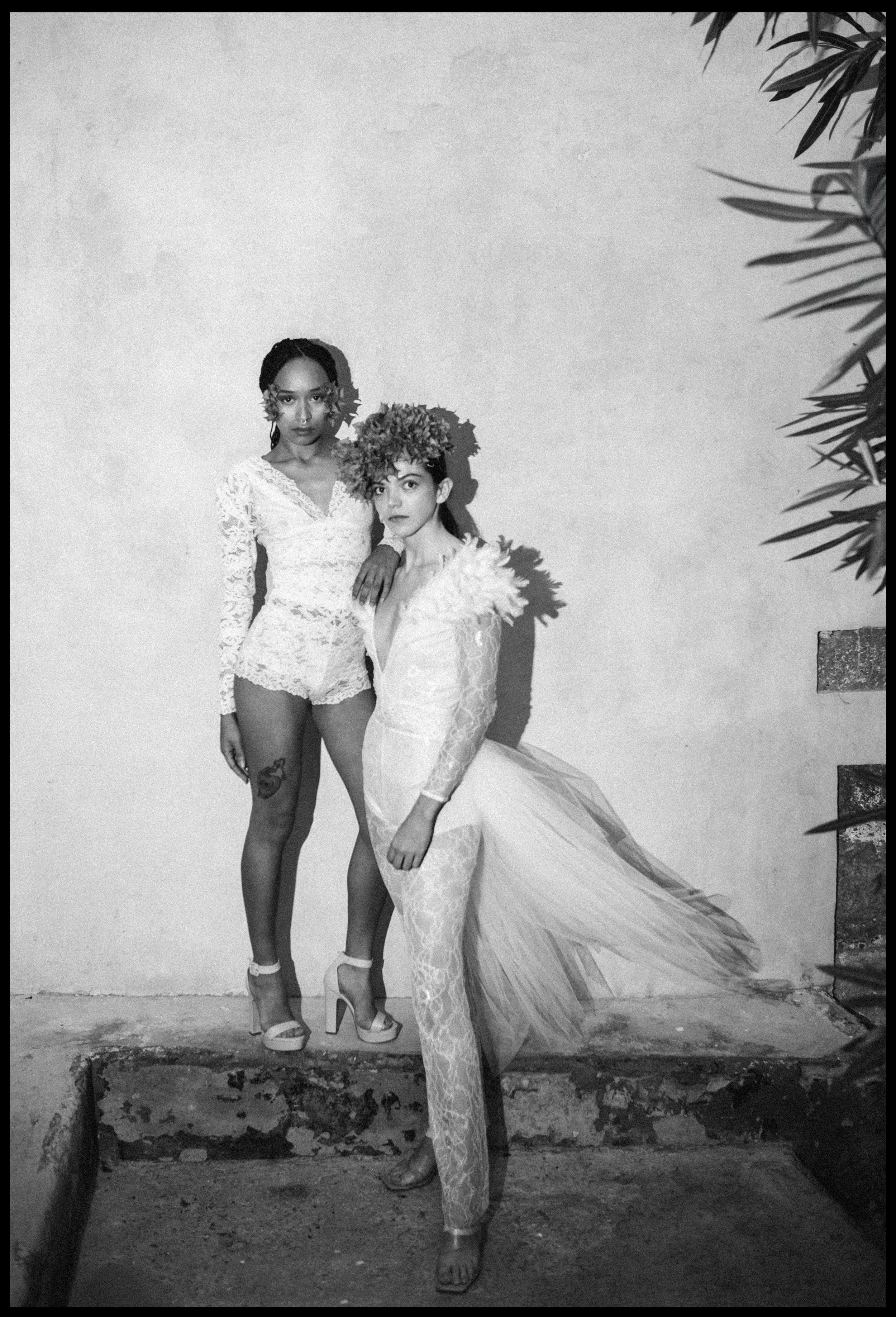 Two women in elaborate lace outfits, one with a floral crown and the other with a feathered collar, standing against a plain wall with a branch partially visible on the right.