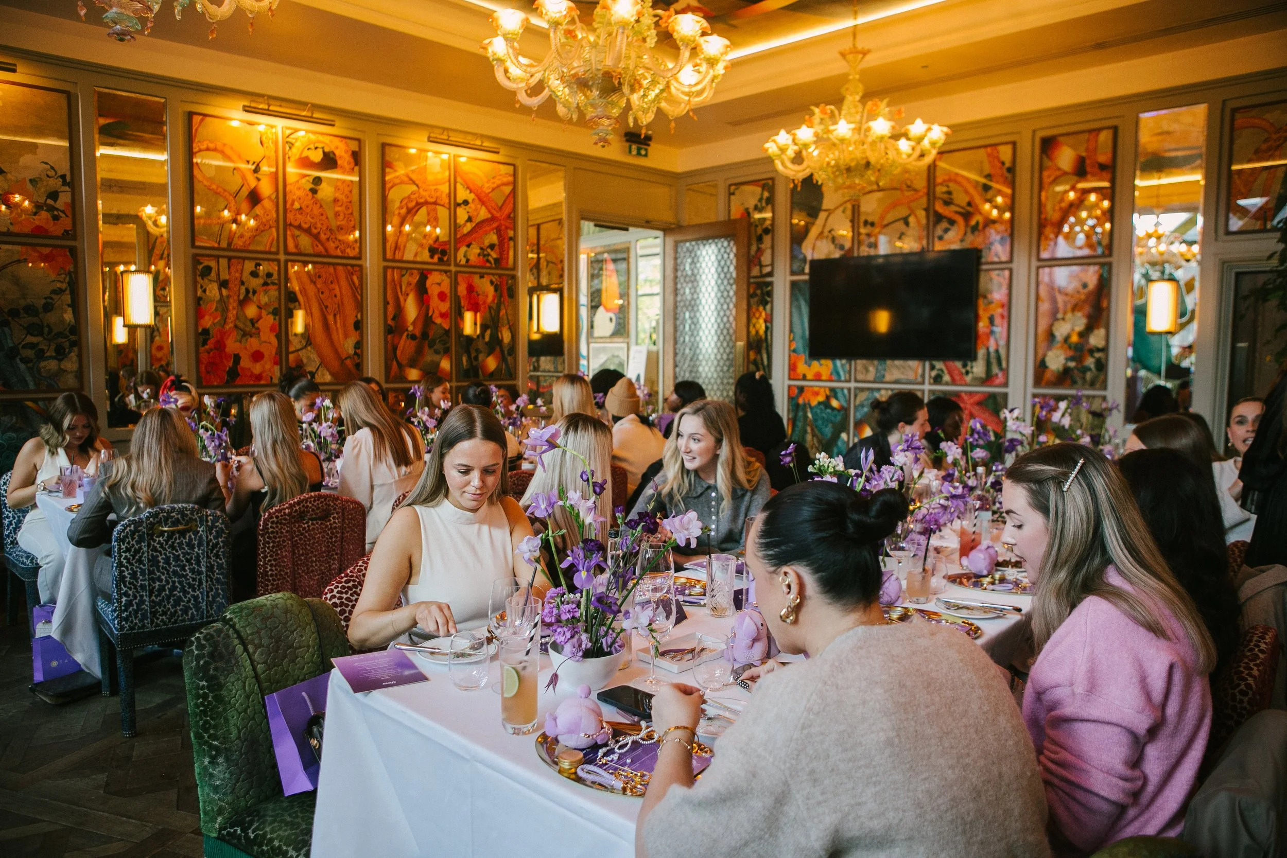A group of women dining at a decorated event with purple flowers, in a room with colorful wall murals and chandeliers.