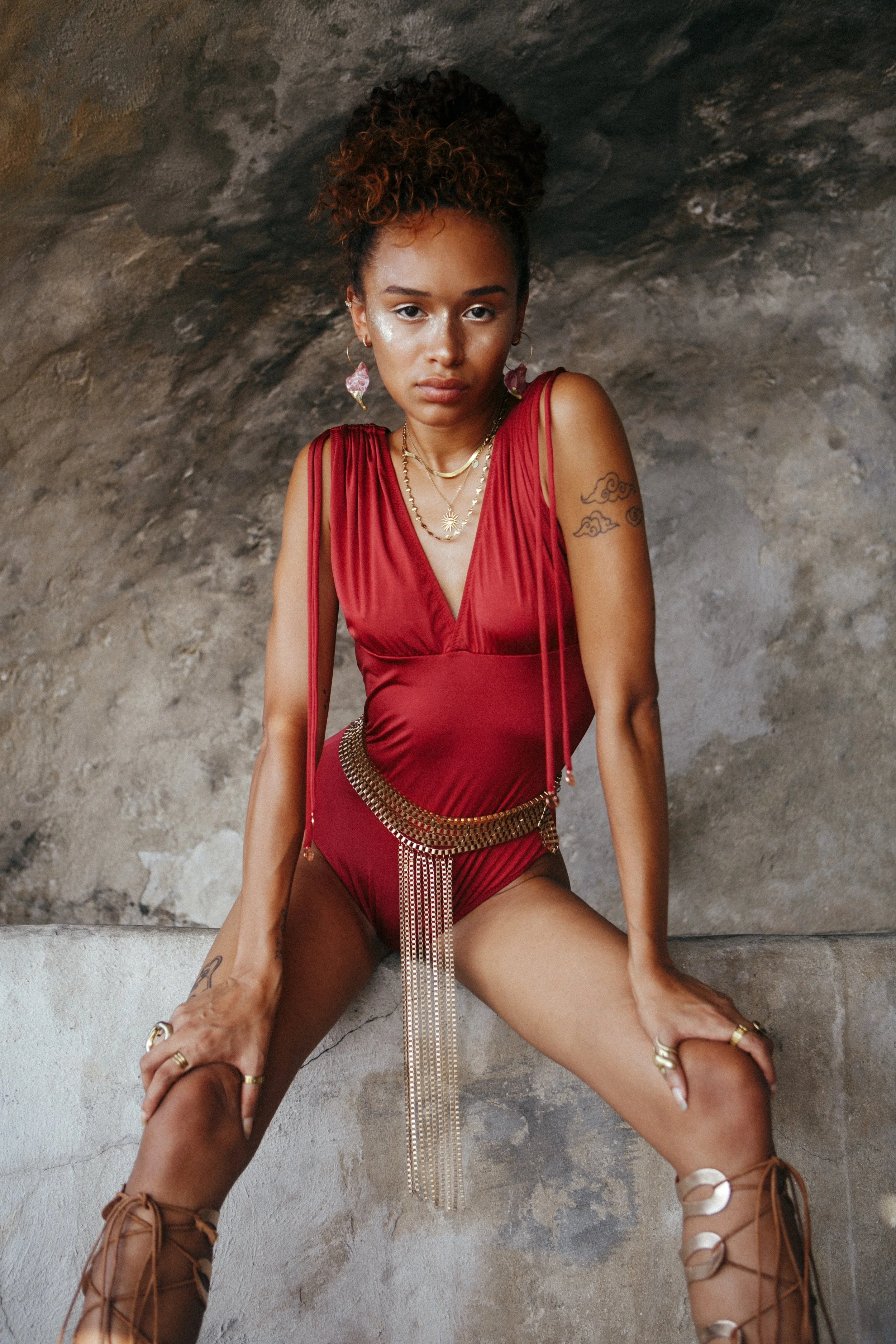 A woman with a serious expression, wearing a red sleeveless outfit, standing against a rocky wall background. She has curly hair styled in a high bun, is adorned with jewelry including necklaces, earrings, and rings, and is wearing tan lace-up boots.