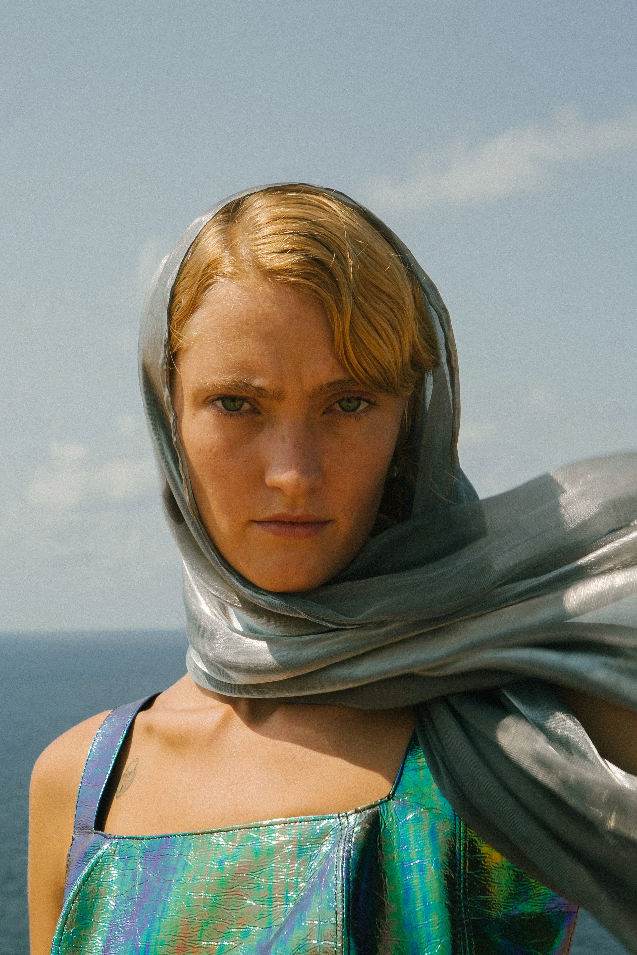 A young woman with blond hair and green eyes, wearing a silver scarf on her head and a holographic dress, standing outdoors near the ocean under a partly cloudy sky.