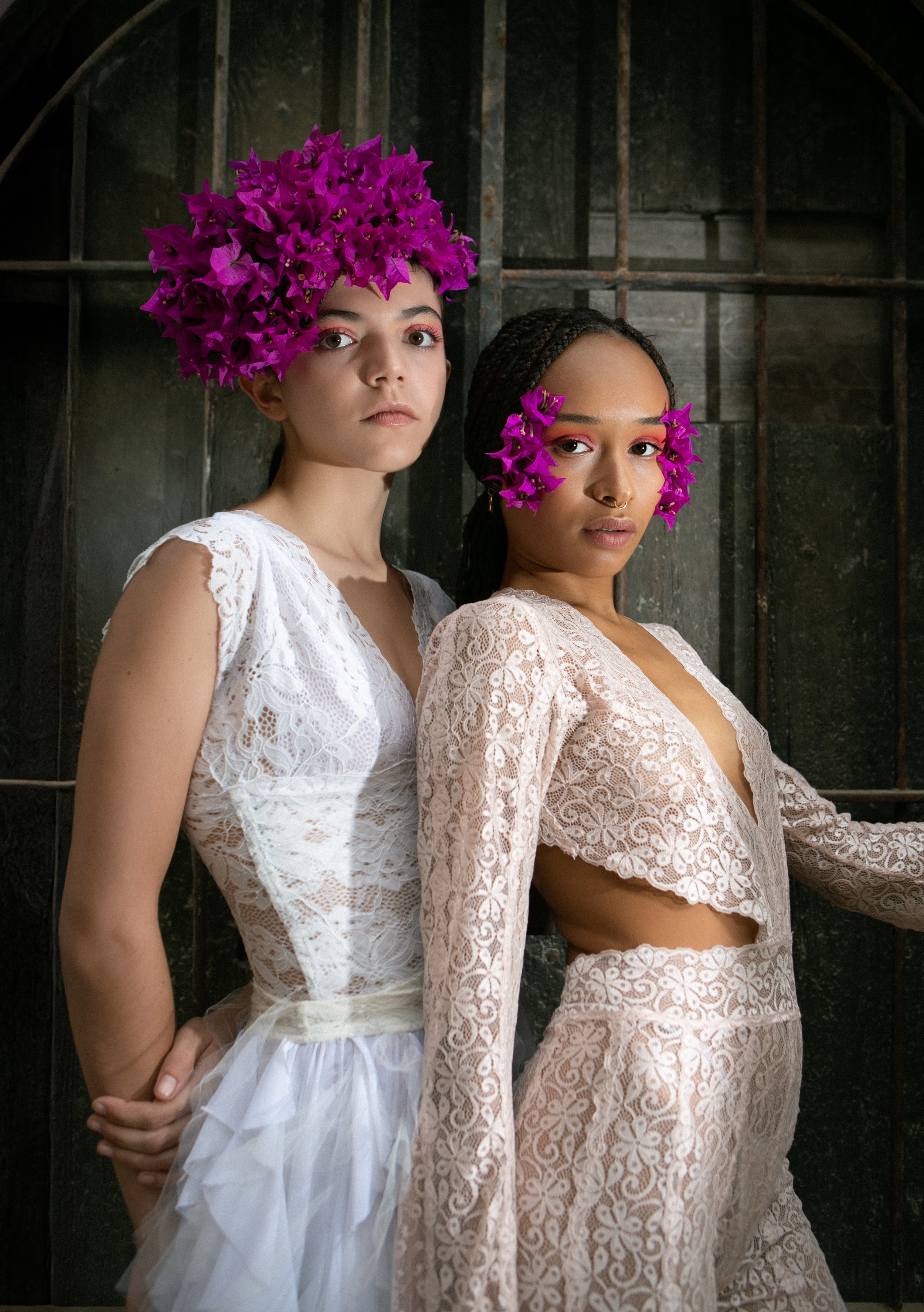 Two women in white lace outfits with pink floral headpieces standing against a dark metal background.