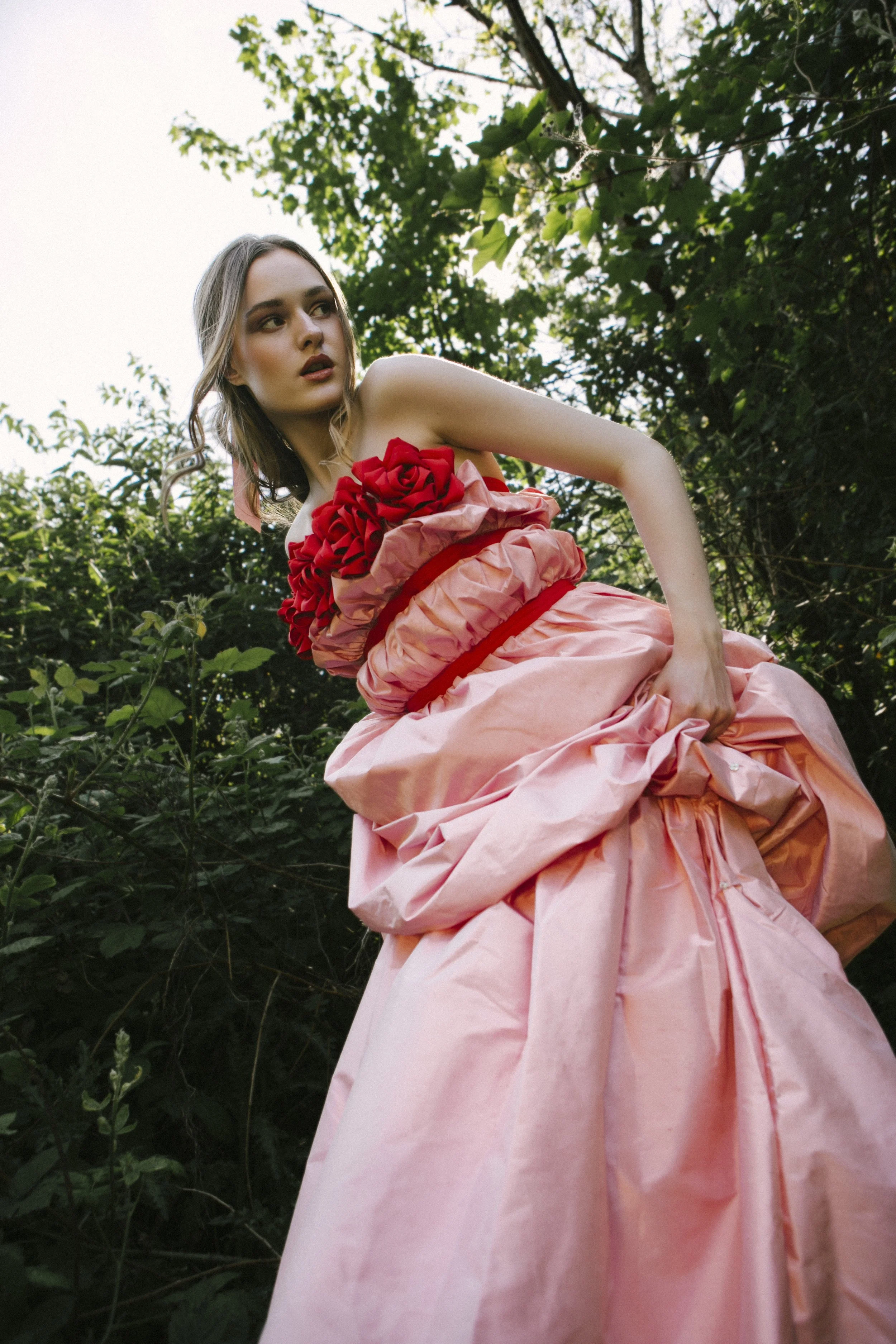 A woman wearing a pink and red floral dress standing outdoors among green foliage.