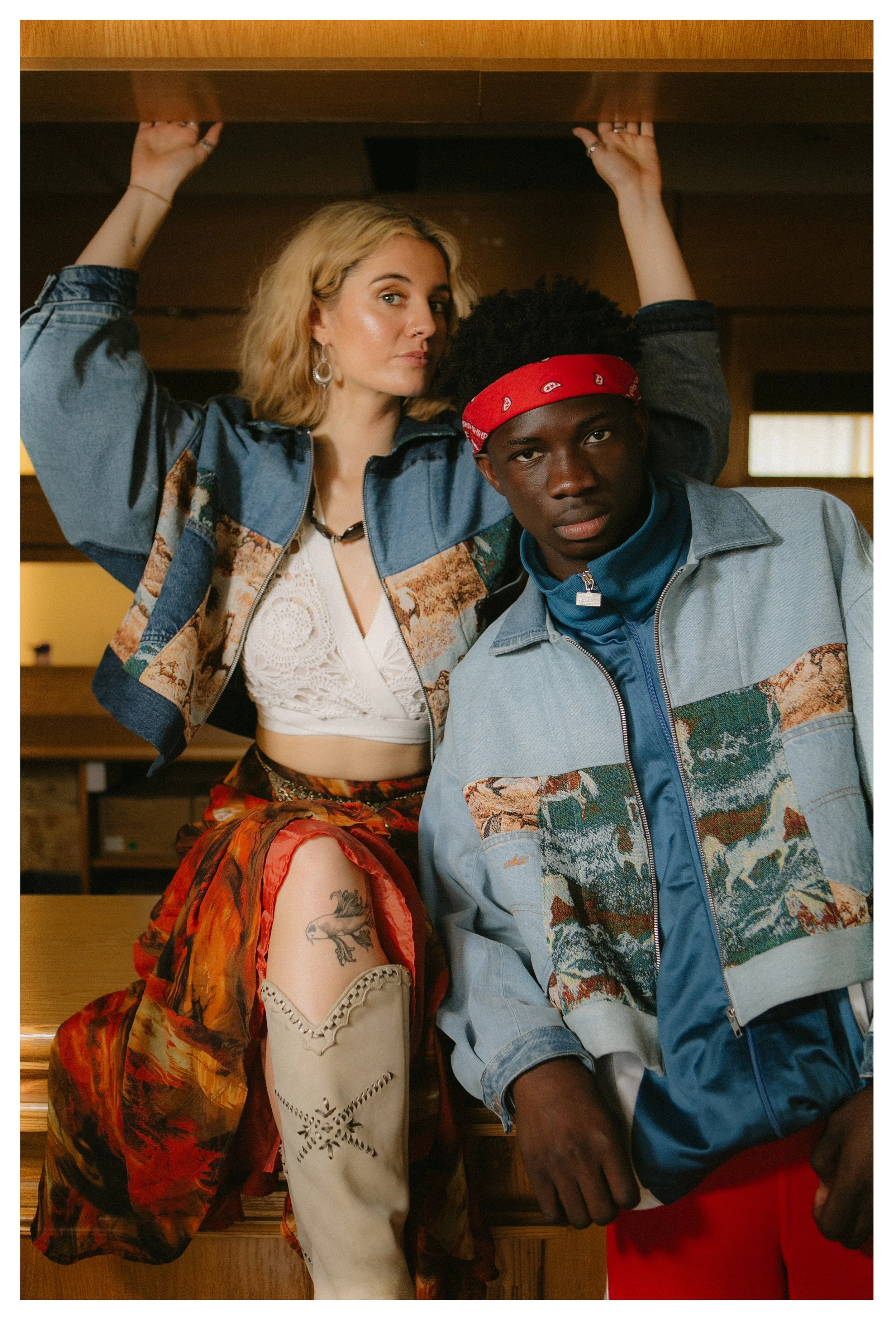 A young woman and man posing indoors. The woman has blonde hair, wears a blue jacket with a scenic print, a white crop top, and a colorful skirt with a thigh tattoo. The man is wearing a matching scenic print jacket, a blue turtleneck, and a red band