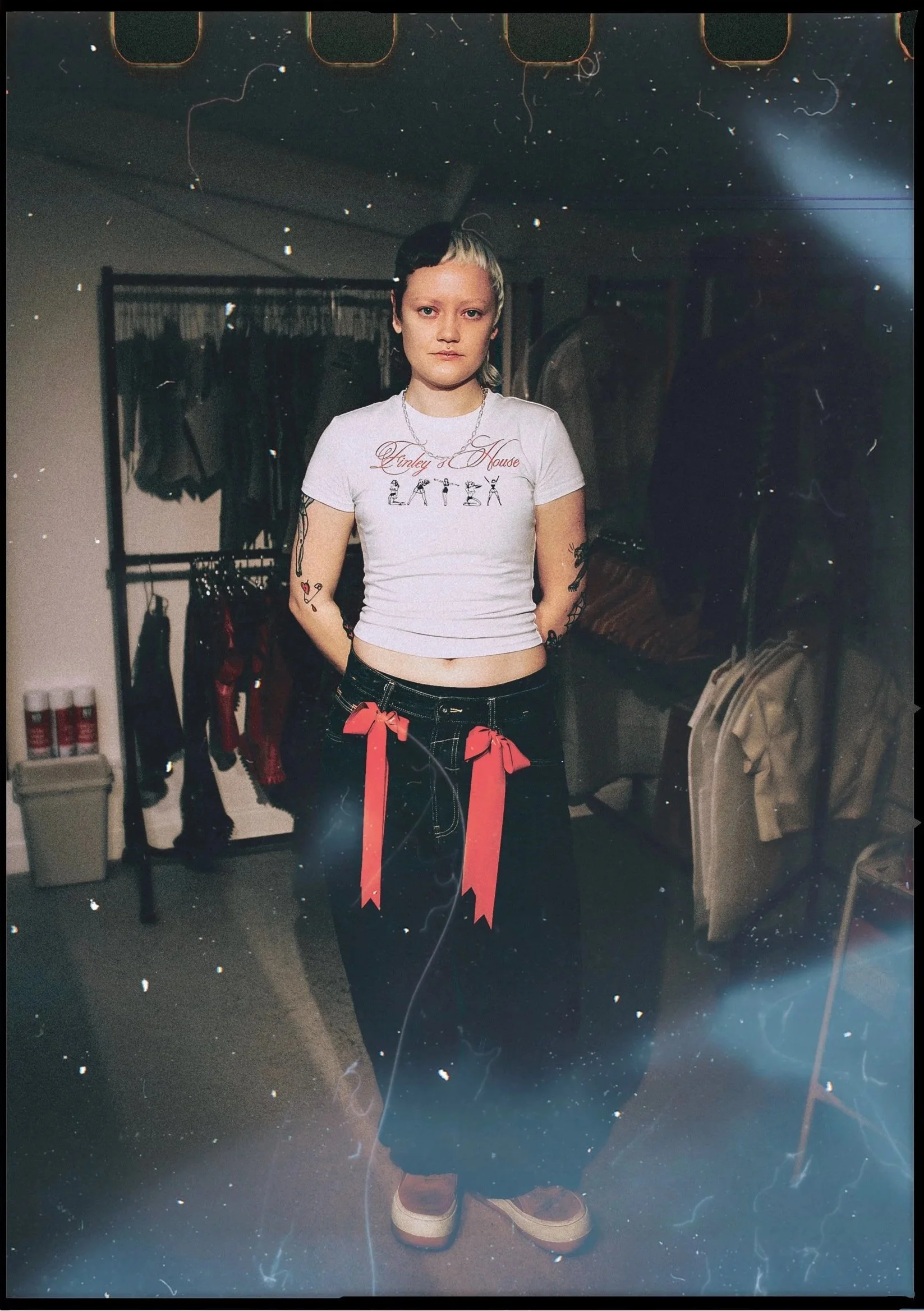 A young woman with tattoos on her arms and short hair, standing in a room with clothing racks and shelves, wearing a white graphic T-shirt, dark jeans with red ribbons hanging from the waist, and pink platform shoes.