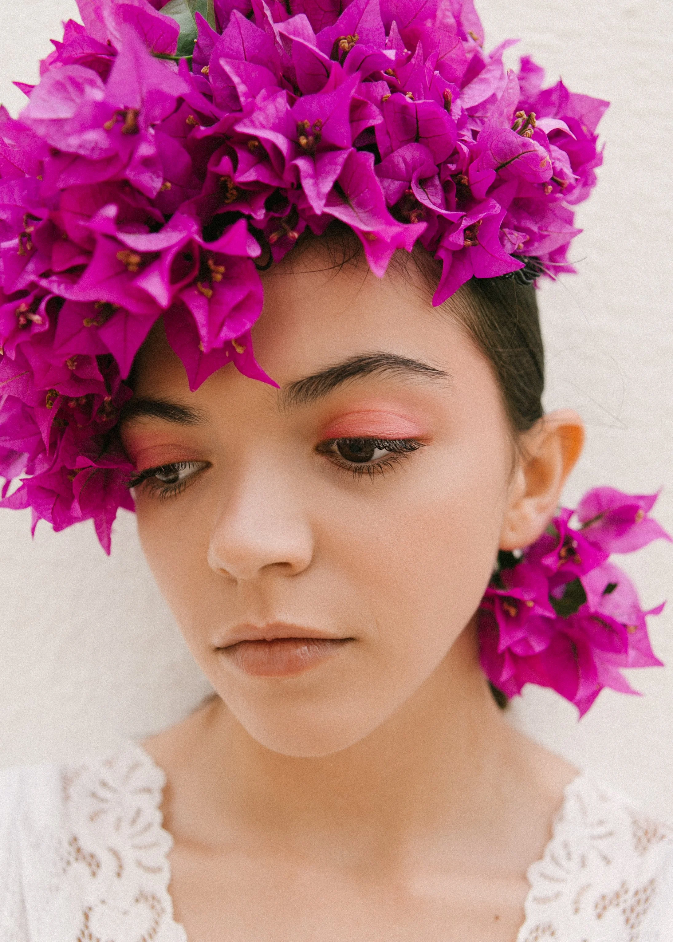 A woman with a floral headpiece made of bright pink bougainvillea flowers, wearing soft makeup with pink eyeshadow and neutral lips, against a plain light background.