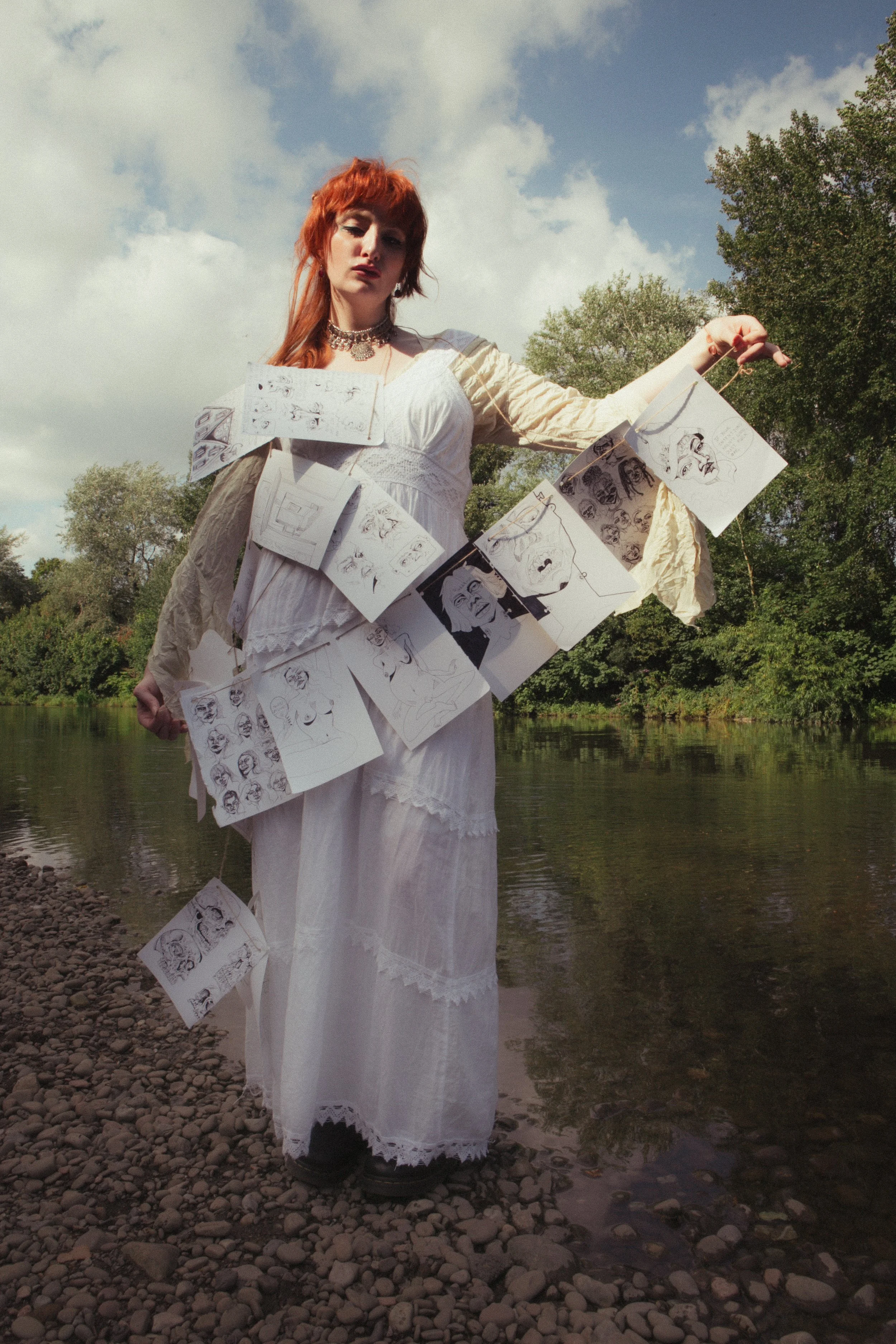 A woman with red hair stands on a rocky riverbank, wearing a white dress and a beige jacket. She has sketches of faces and comic-style illustrations hanging around her body, displayed on strings.