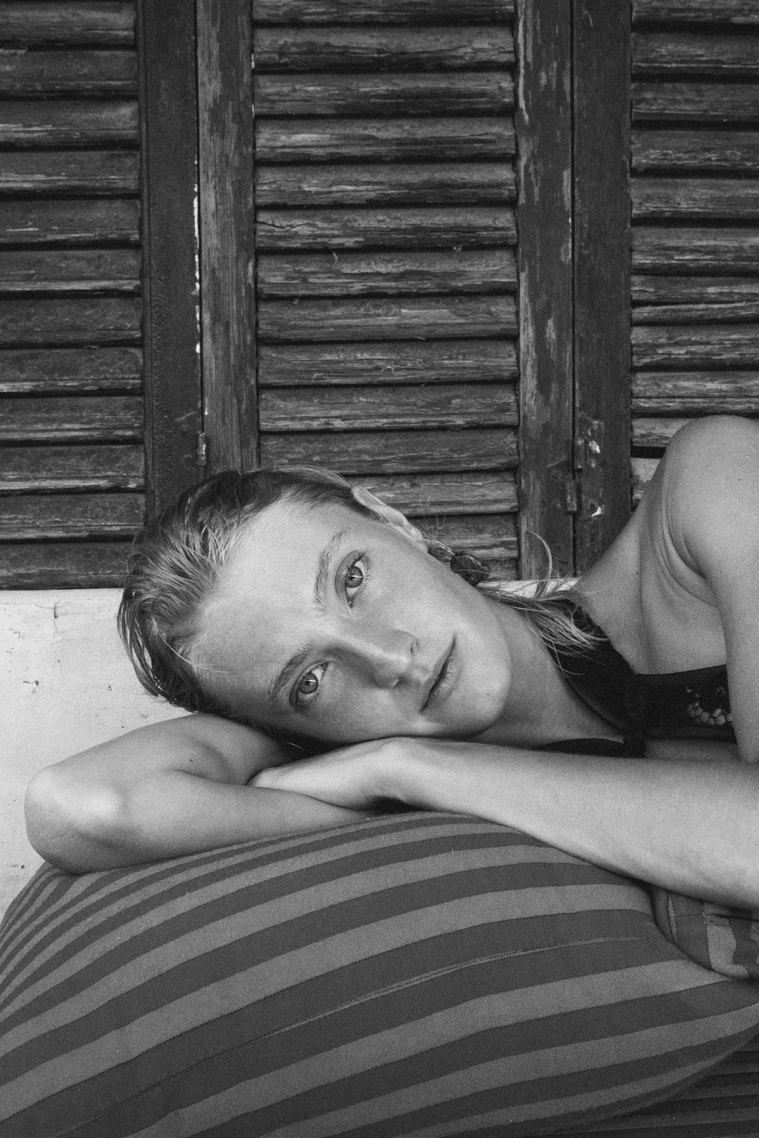 A young woman with wet hair lying on a striped pillow, resting her head on her arm, looking directly at the camera, with a wooden shutter background.