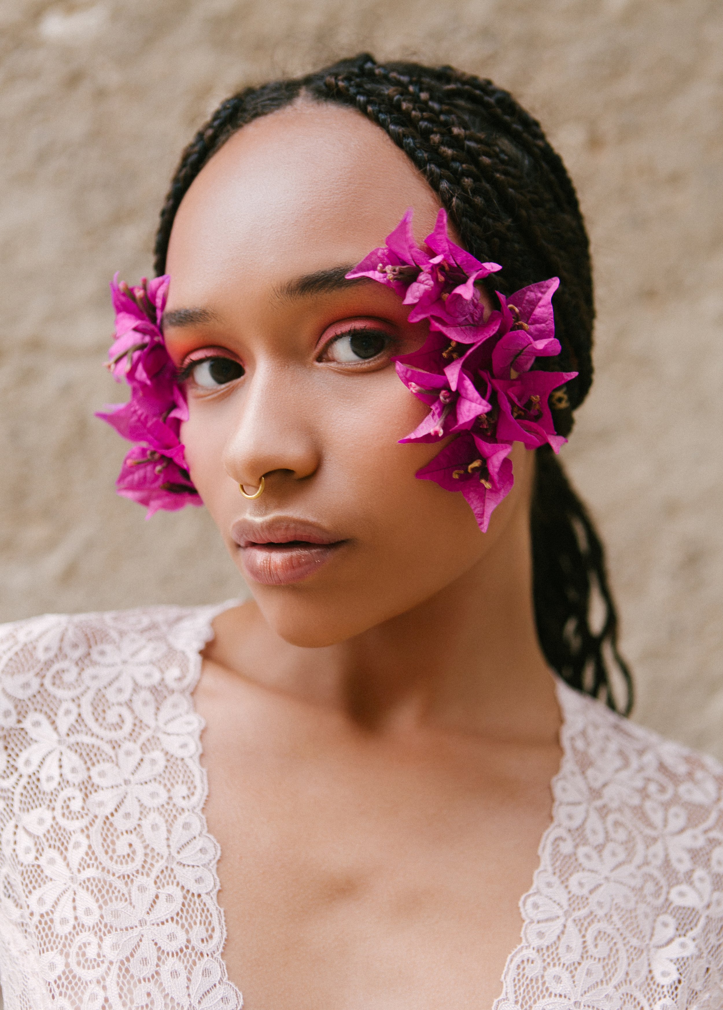 A woman with dark braids and a septum piercing, with pink flowers around her eyes, wearing a white lace top, against a beige background.