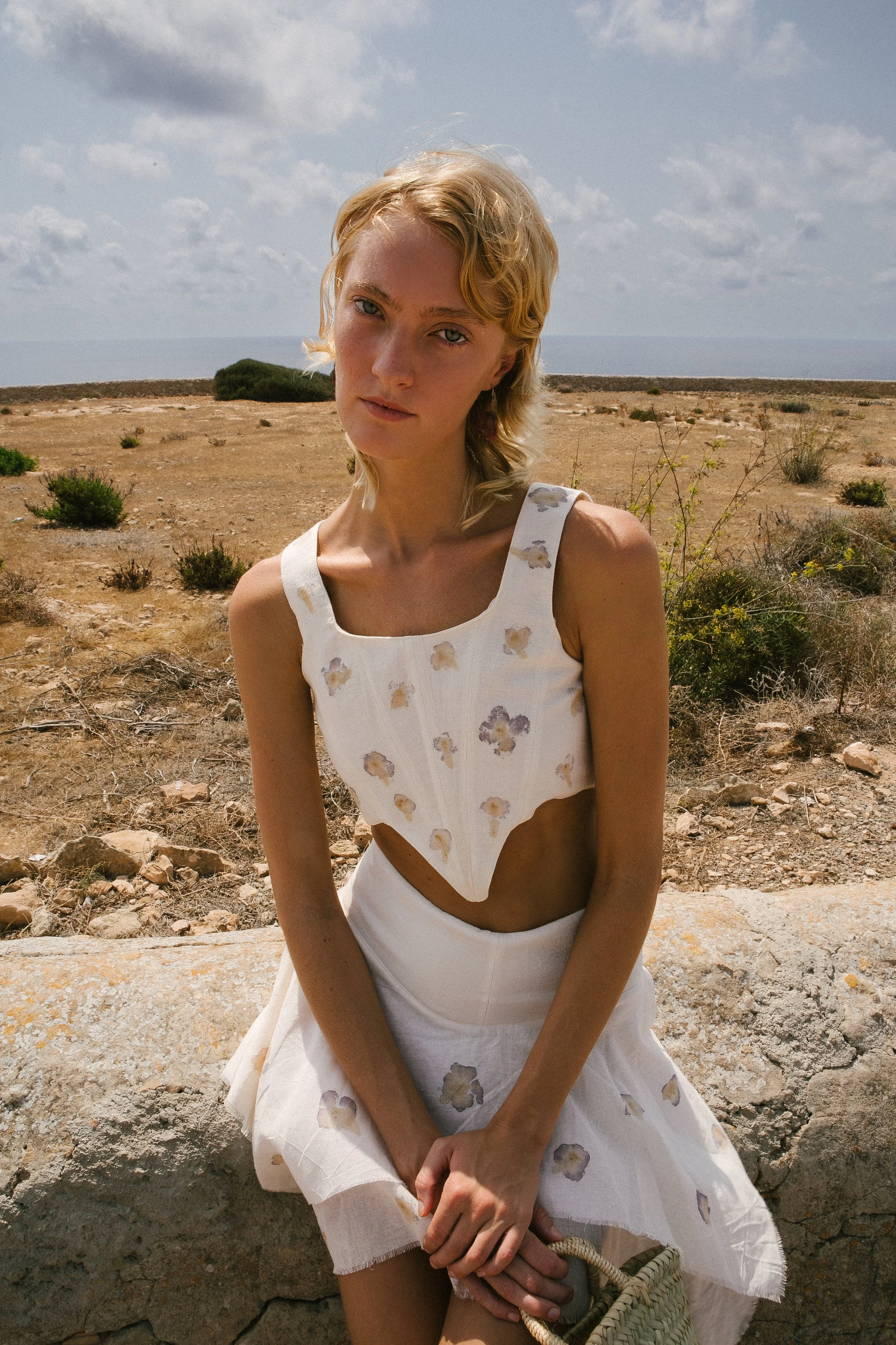 A young woman sitting outdoors in a dry, desert-like landscape with sparse shrubs, wearing a white, sleeveless dress with floral patterns and cutouts, holding a small woven bag, and looking at the camera with a neutral expression.
