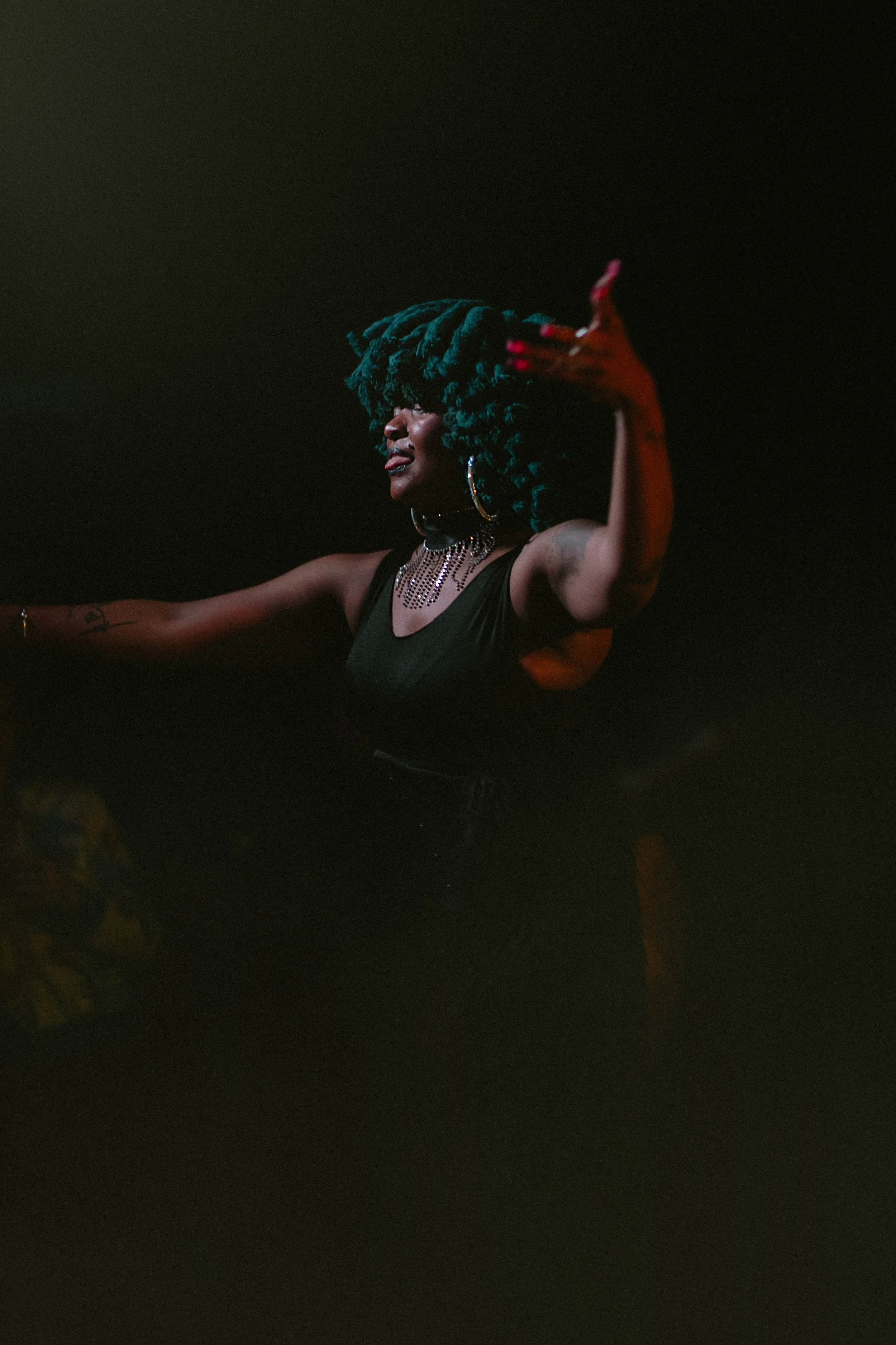A woman with large, dark green dreadlocks, wearing a black sleeveless top, hoop earrings, and layered necklaces, appears to be speaking or performing on stage with a dark background.