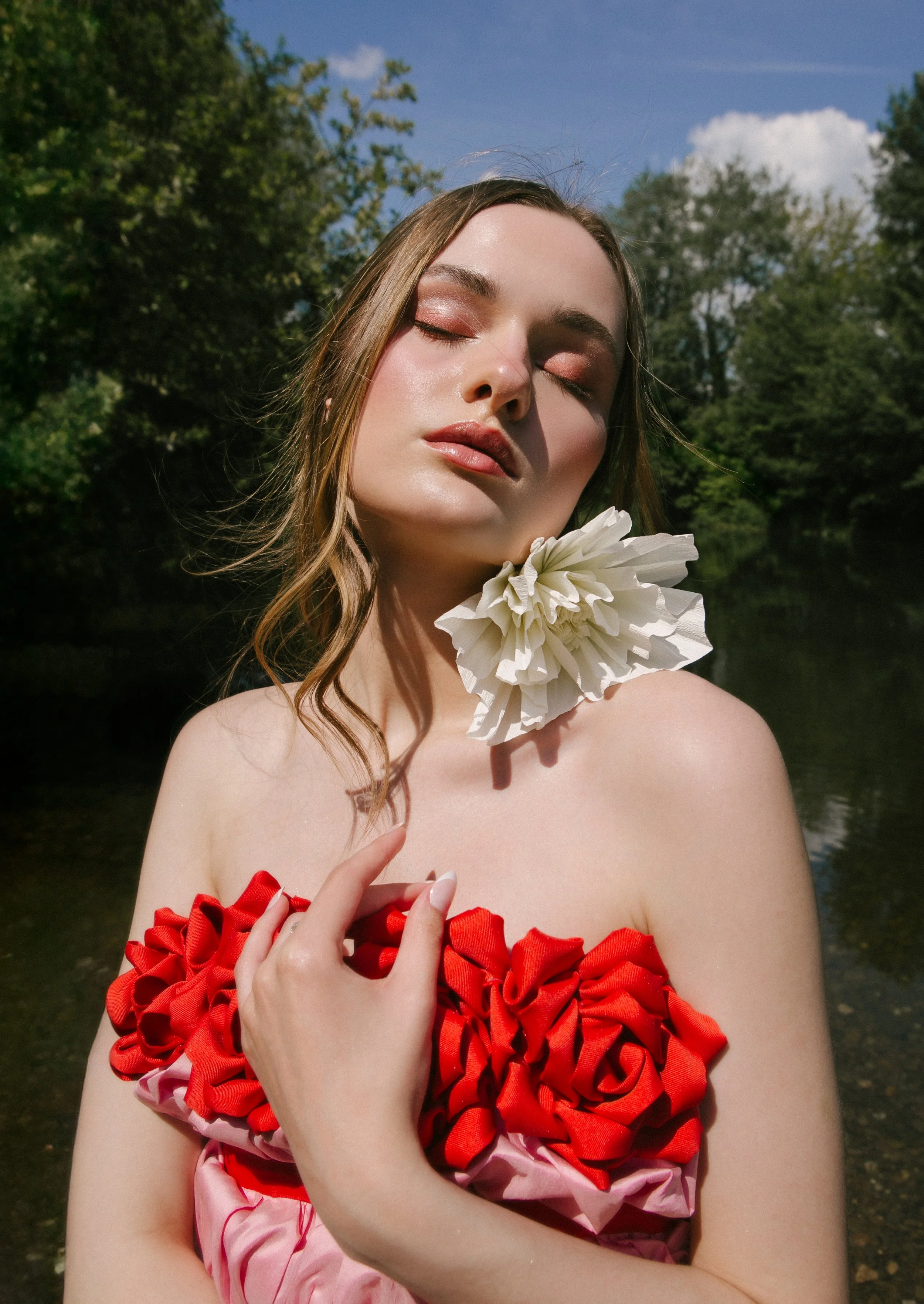 A young woman with closed eyes, light makeup, and long brown hair stands outdoors by a body of water. She wears a strapless dress made of red and pink fabric roses and has a large white flower around her neck. The background features trees and a part