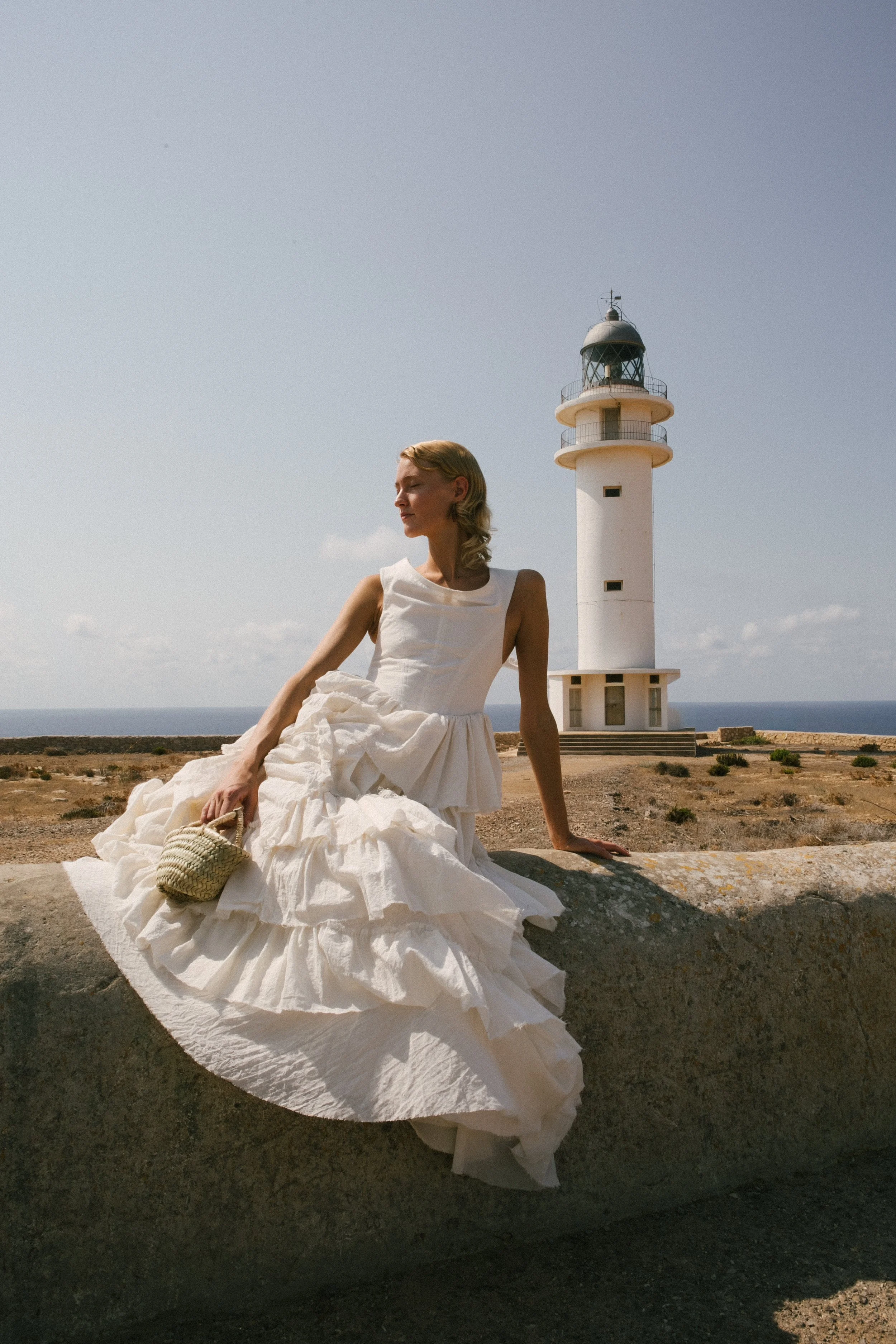 A woman in a white dress sitting on a large rock, holding a straw basket, with a lighthouse and ocean in the background.