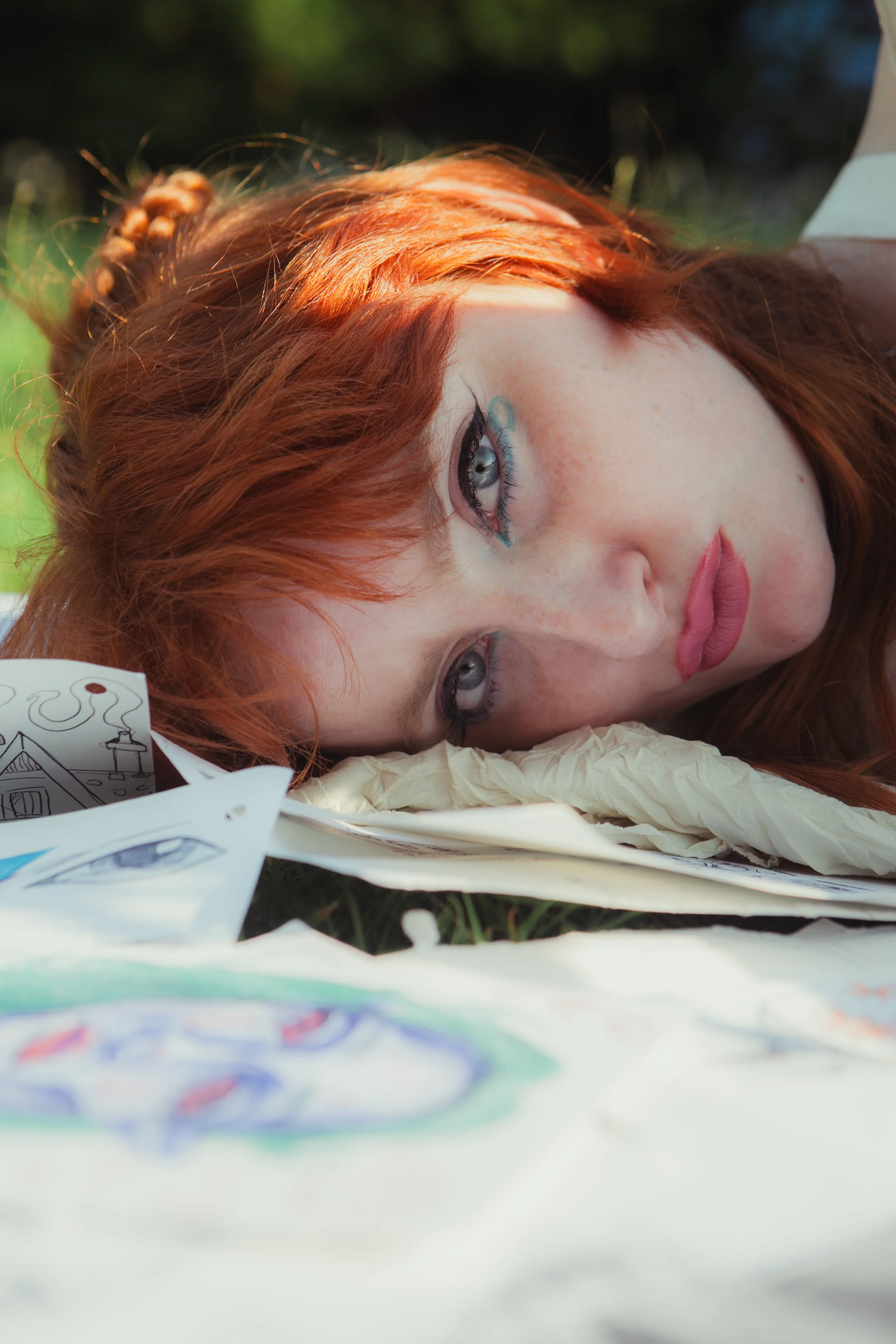A red-haired woman with blue eyes lying on the ground surrounded by drawings and sketches.