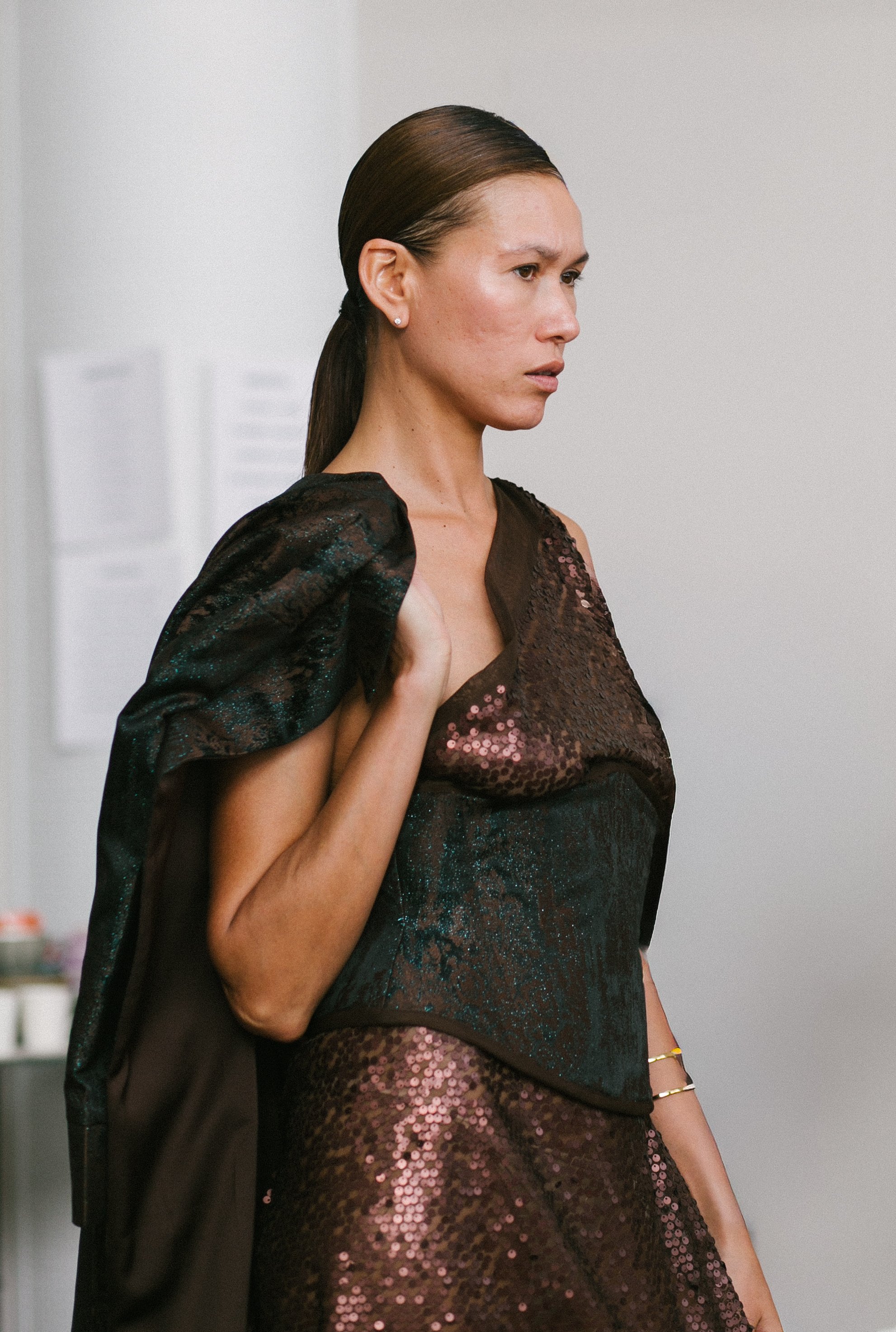 A woman with brown hair tied back, wearing a shiny black and pink sequined dress, holding a black jacket over her shoulder, standing indoors against a plain light-colored background.