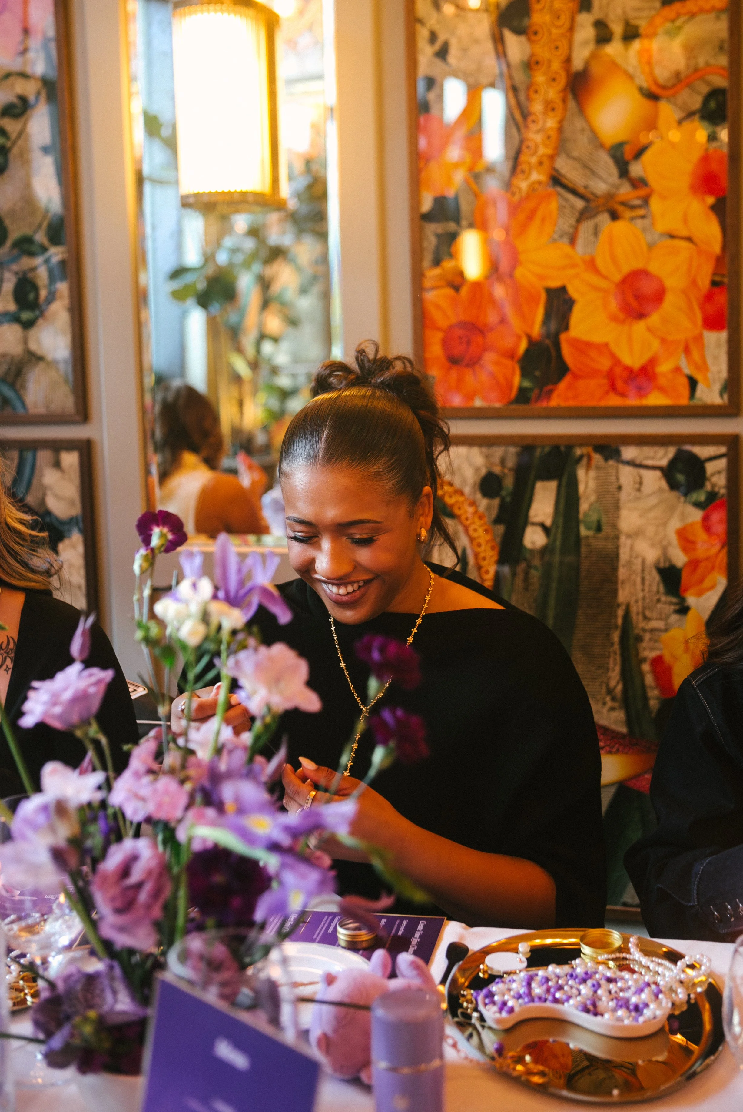 A woman with dark hair in a ponytail, wearing a black top and jewelry, smiling while arranging purple and pink flowers at a decorated table in a restaurant with floral artwork on the walls.