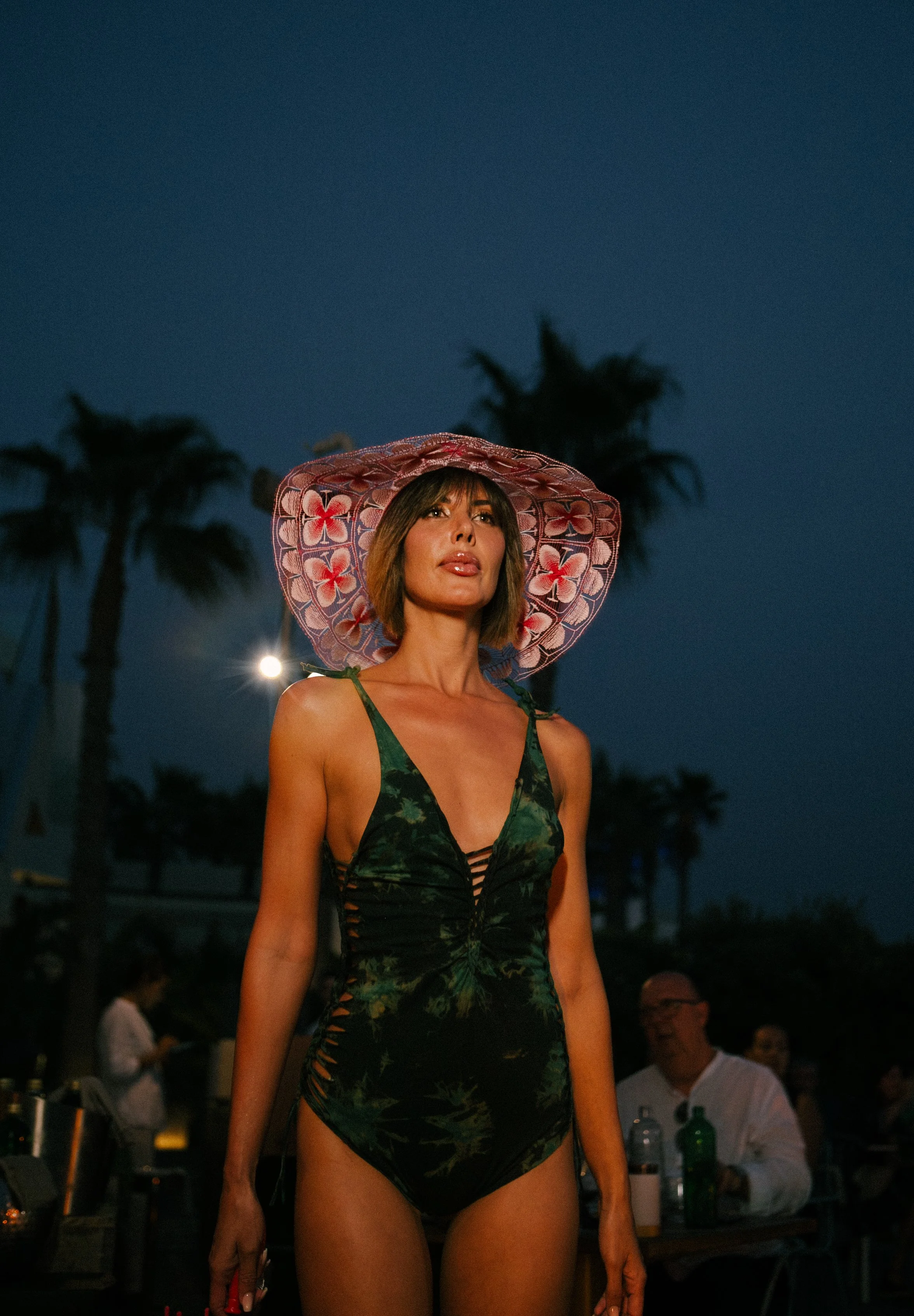 A woman wearing a large pink floral sunhat and a dark green swimsuit with a tropical print standing outdoors at night with palm trees in the background.